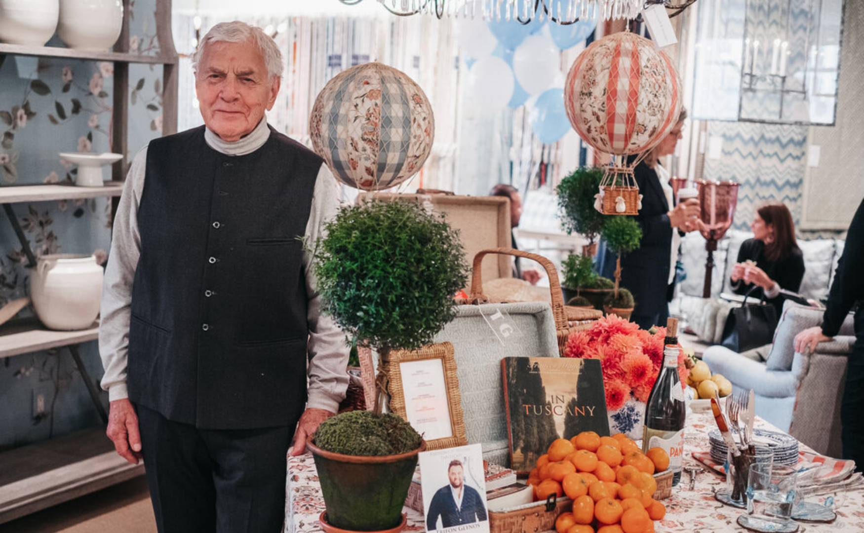 An elderly man with white hair wearing a black vest and white shirt standing at a table with plants, oranges, a bottle of wine, and decorative hot air balloons at an indoor market or shop.
