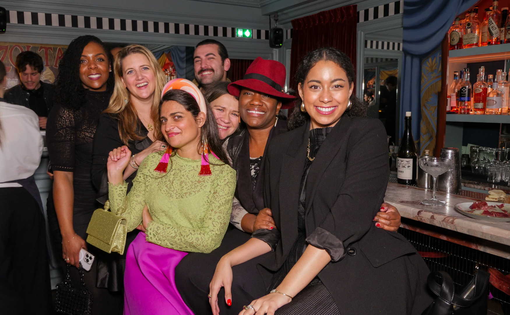 Group of women smiling and posing together at a social gathering in a bar or restaurant, with shelves of liquor bottles in the background.