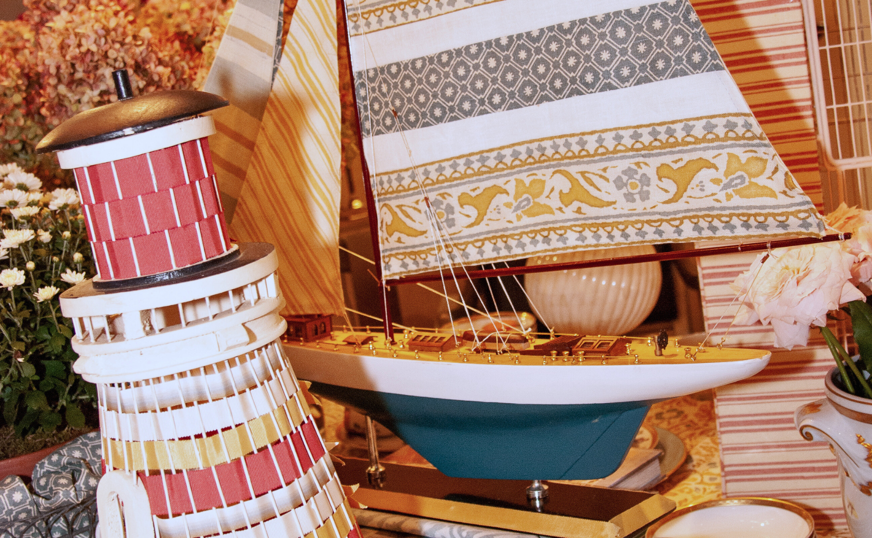 Detailed model sailboat with a white hull, blue bottom, and ornate sail, surrounded by decorative objects including a brightly colored woven lamp and floral arrangements.