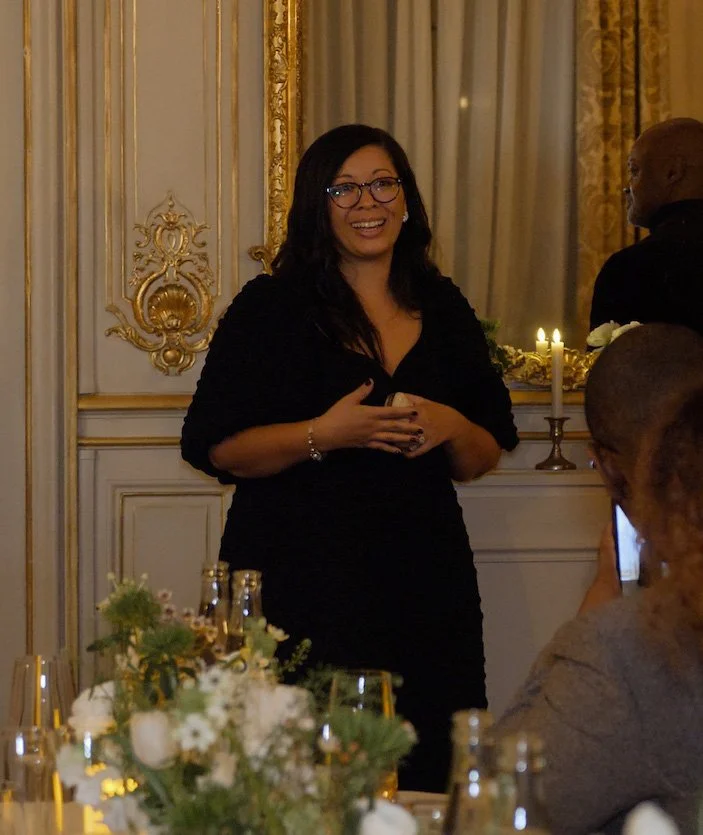A woman with dark hair, glasses, wearing a black dress, smiling and speaking at a formal event in an ornate room with gold decorations and candles, surrounded by seated guests.