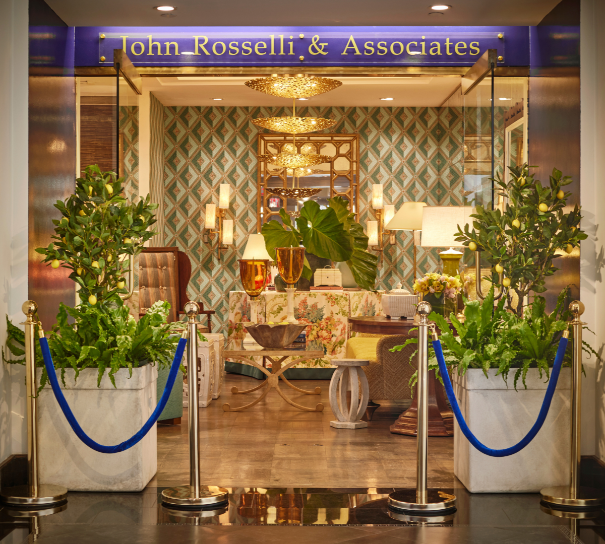Elegant hotel lobby with plants, gold accents, and vintage furniture, viewed through glass doors with the sign "John Rosselli & Associates" above.