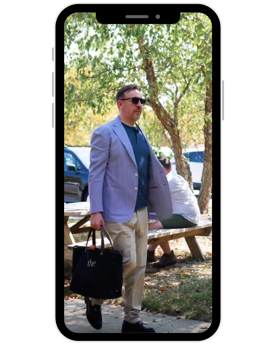 A man wearing sunglasses, a light blue blazer, a dark shirt, and khaki pants, is walking outdoors carrying a large black tote bag. There are trees, a wooden picnic table, and a man sitting in the background.