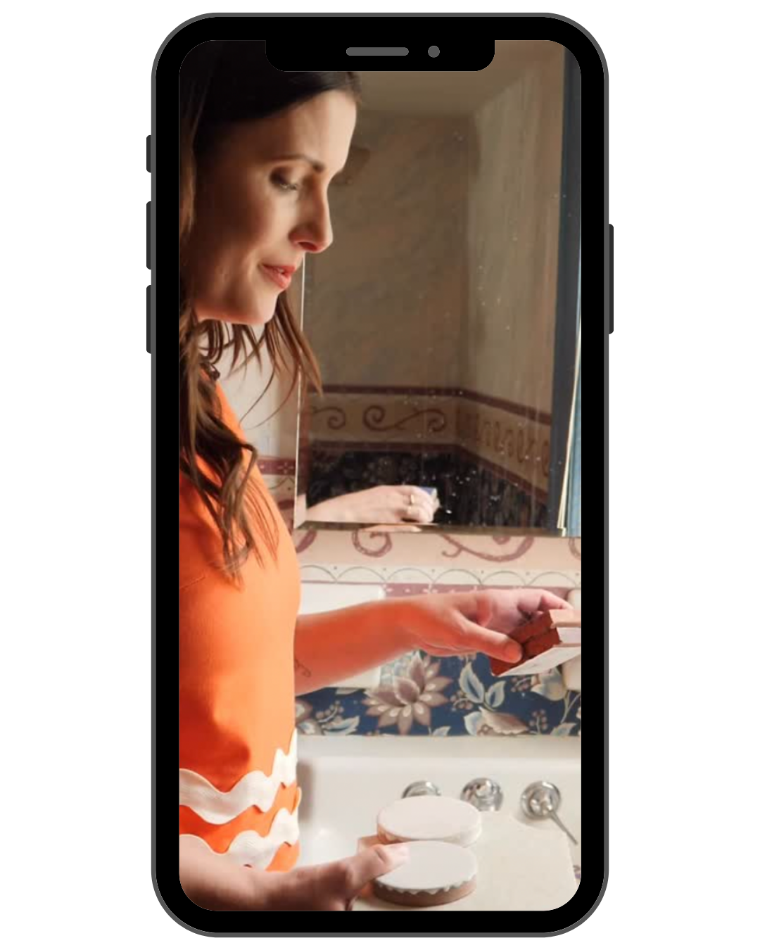 A woman looking into a mirror in a bathroom, holding two bottles of skincare or cosmetic products.