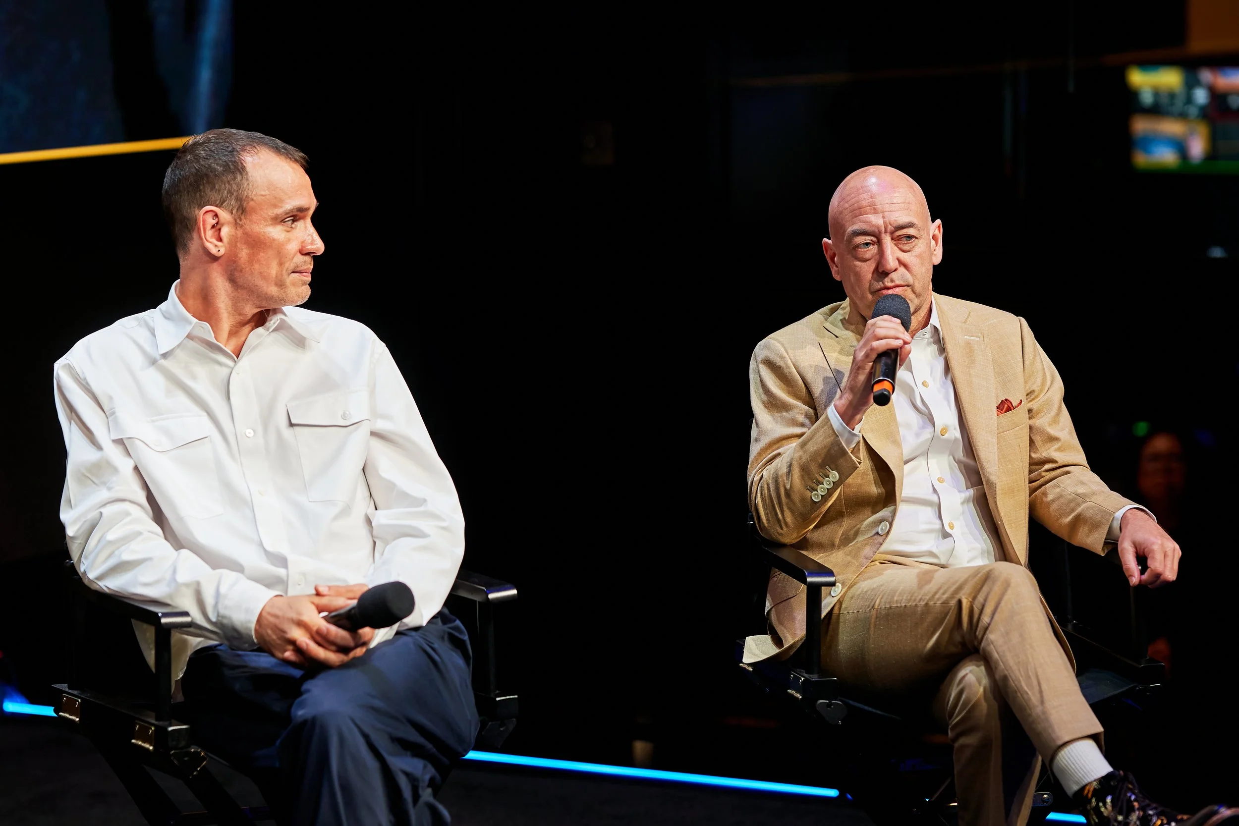 Two men sitting on stage in black chairs, one holding a microphone, during a panel discussion or interview.