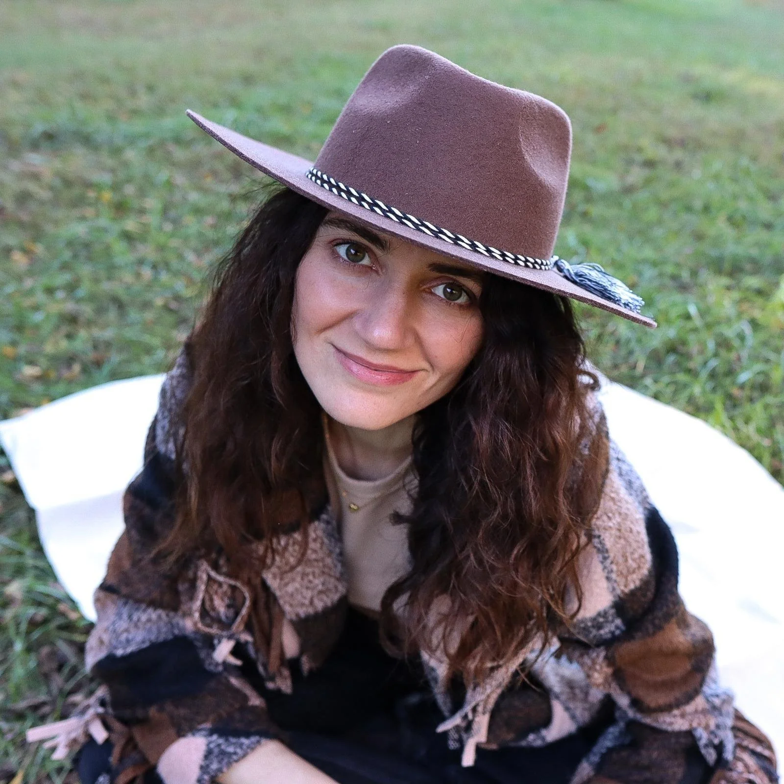 Woman wearing a brown wide-brimmed hat with decorative band, smiling, outdoors on grass.