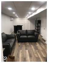 Living room with black leather sofas, wooden floors, and white walls illuminated by ceiling lights.