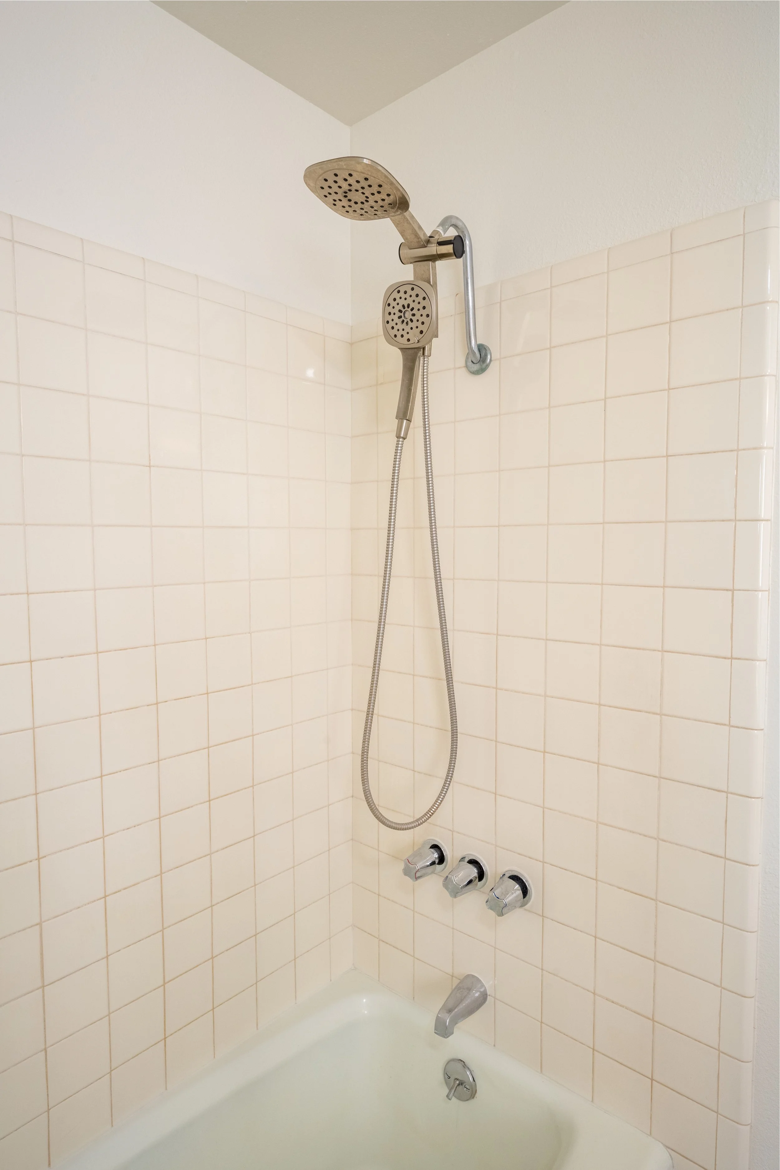 Bathroom shower with beige tiled walls, a handheld showerhead, a fixed showerhead, and three control knobs.