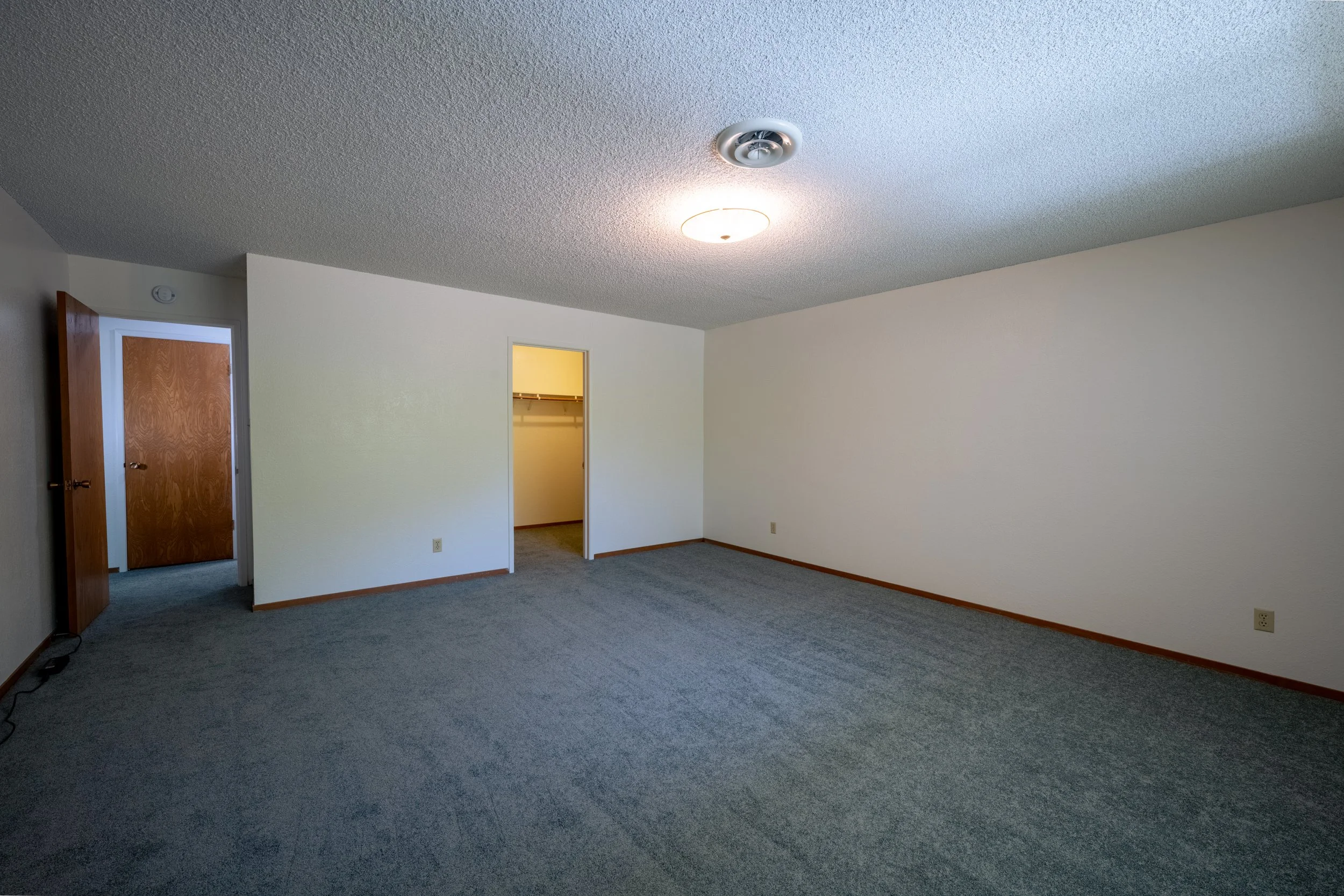 Apt. F Canary Apartments in Calistoga, CA.  Empty room with white walls, gray carpet, closets, and wooden doors, with ceiling light and vent.
