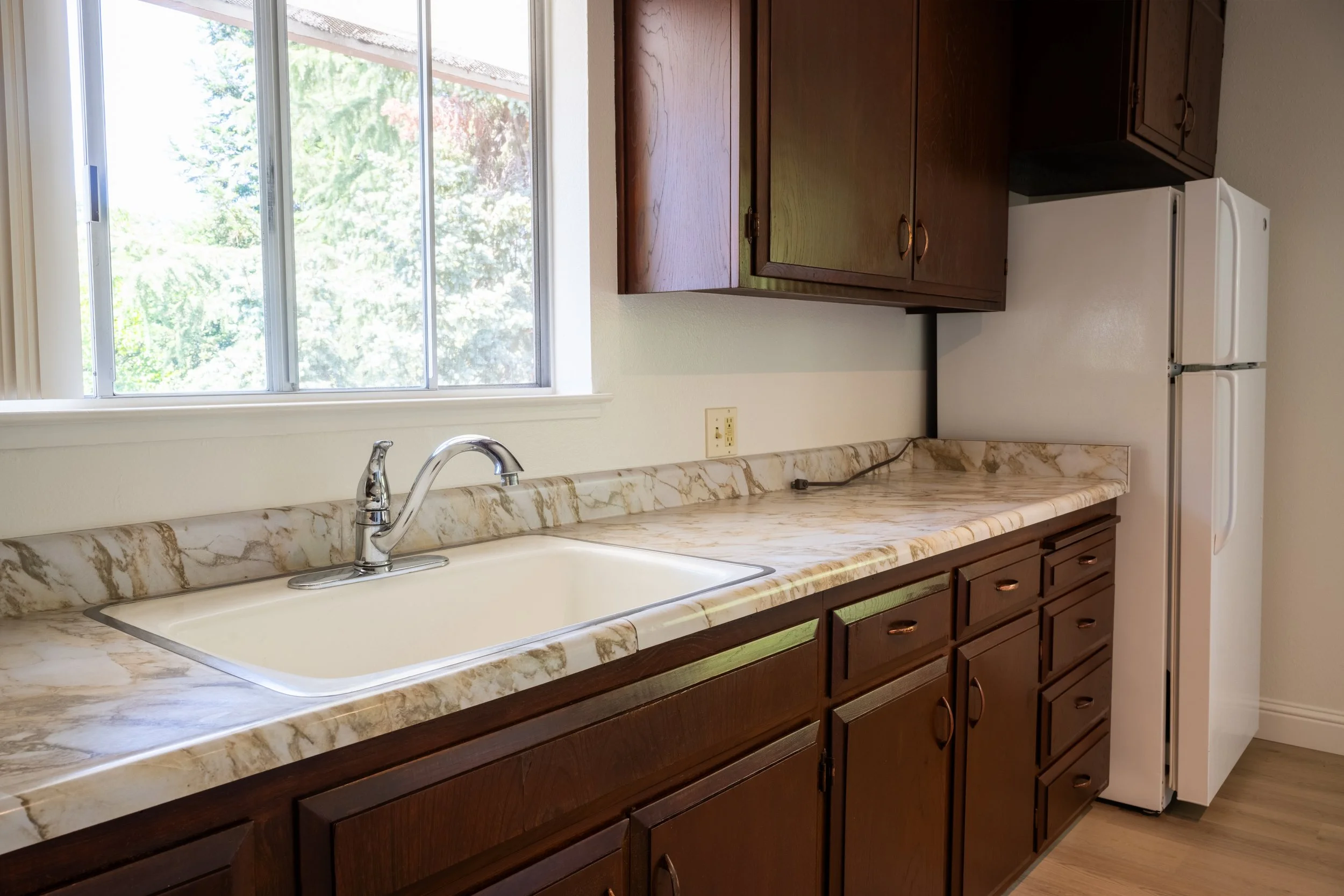 Apt. F: Canary Apartments in Calistoga, CA.  Kitchen with brown wooden cabinets, marble countertop, white sink, chrome faucet, refrigerator, and window overlooking trees.