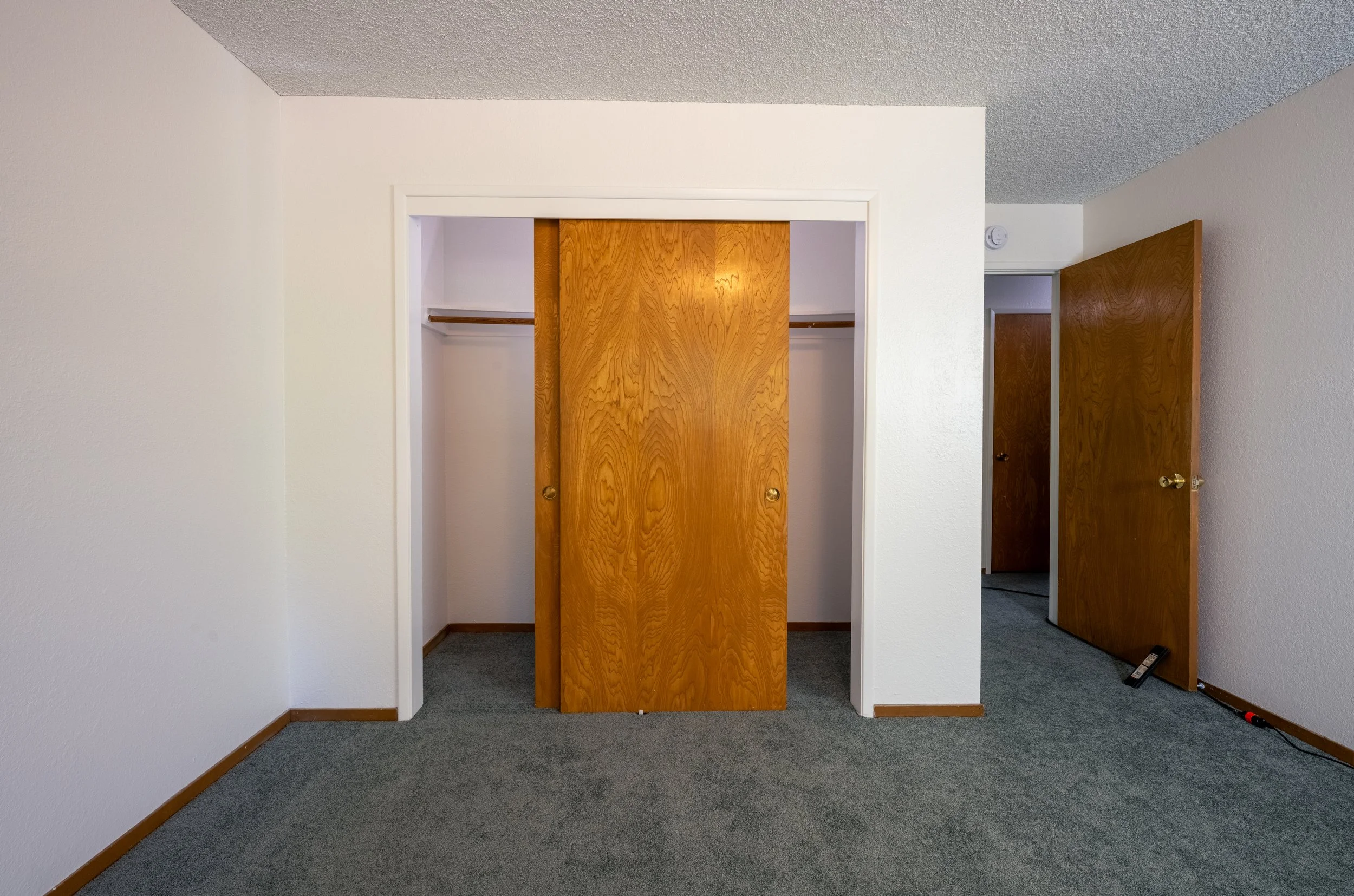 Apt. F Canary Apartments in Calistoga, CA. Empty room with white walls, gray carpet, wooden closet doors, and another door open leading to a hallway.