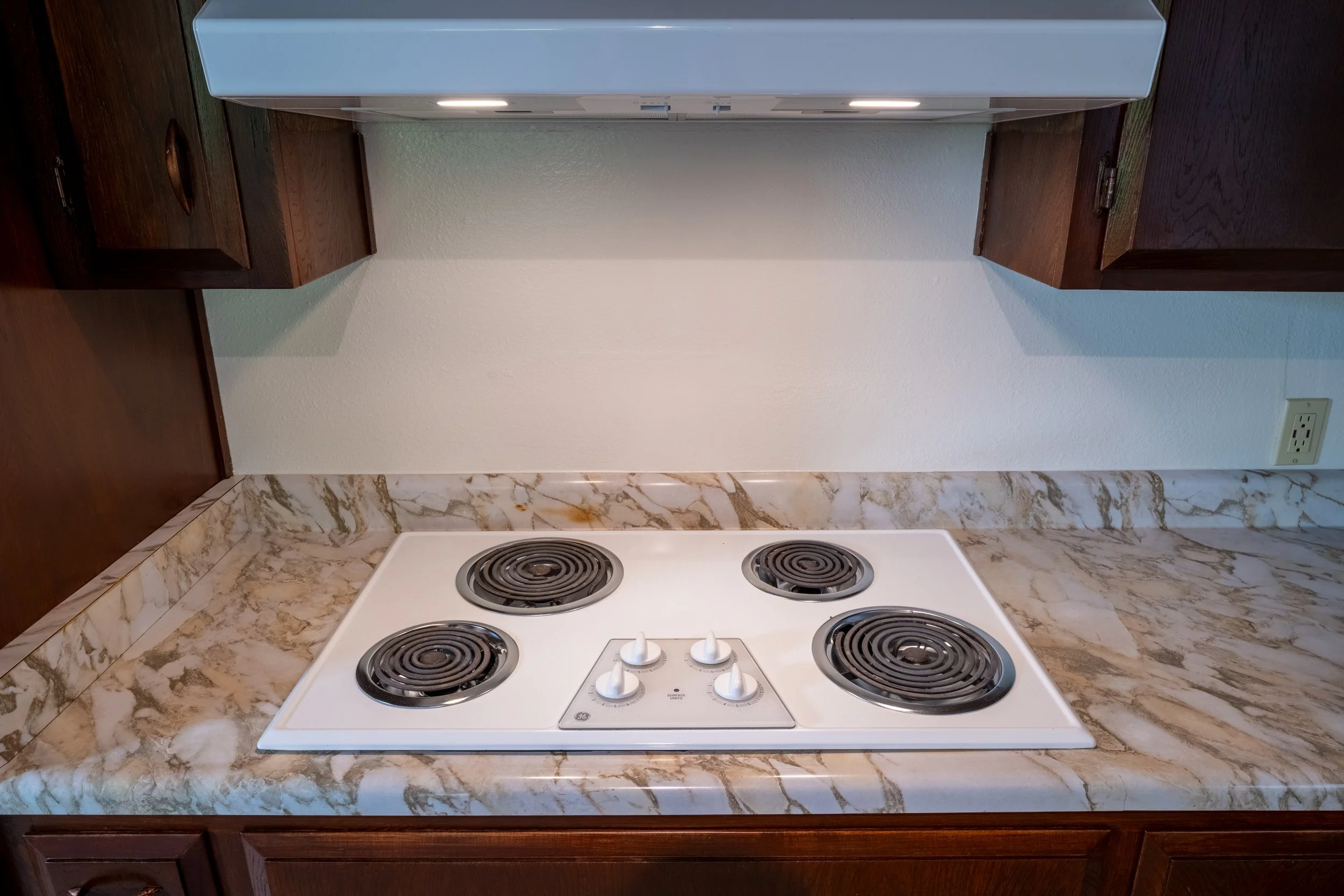 Apt. F: Canary Apartments in Calistoga, CA.   White electric stovetop with four coil burners and knobs, surrounded by a marble countertop and dark wooden cabinets.