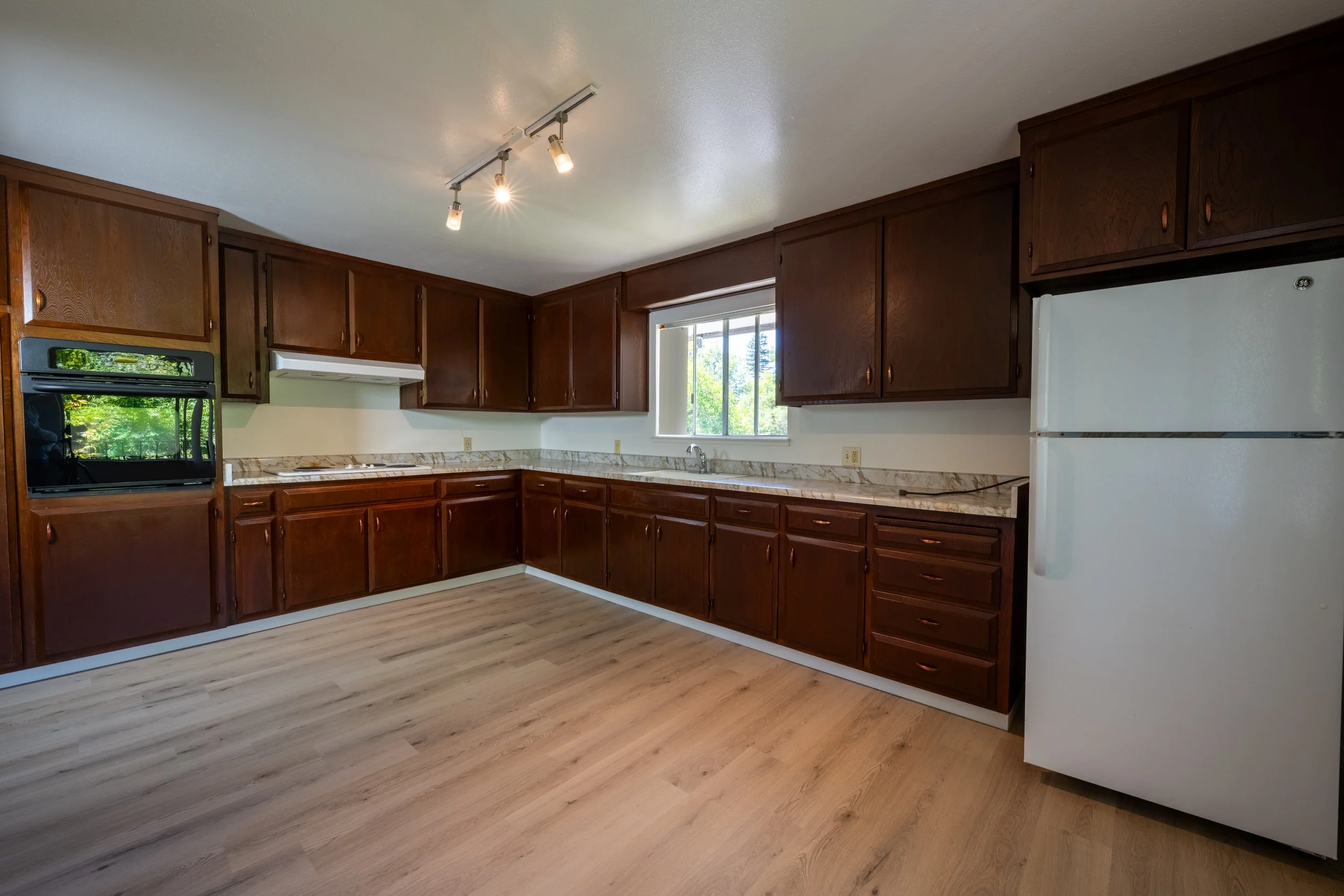 Apt. F: Canary Apartments in Calistoga, CA. Kitchen with wooden cabinets, white refrigerator, marble countertop, and a window showing green trees outside.