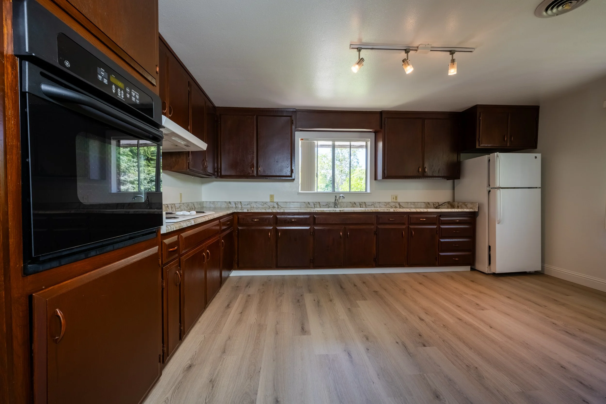 Apt. F: Canary Apartments in Calistoga, CA.  Kitchen with wooden cabinets, white refrigerator, black microwave oven, marble countertop, window above the sink, and wooden flooring.