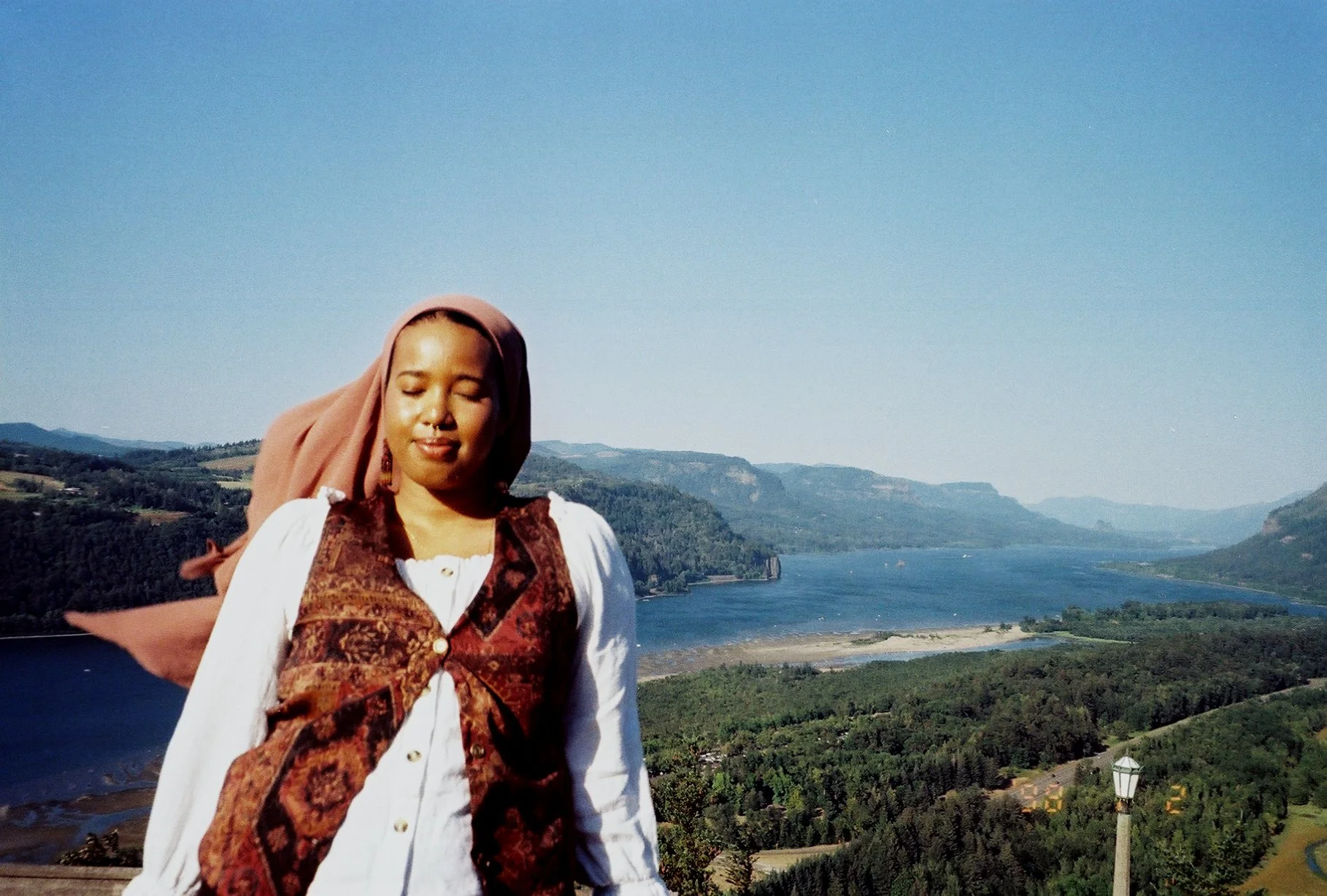 A woman standing outdoors with a river and forested hills in the background on a clear day.