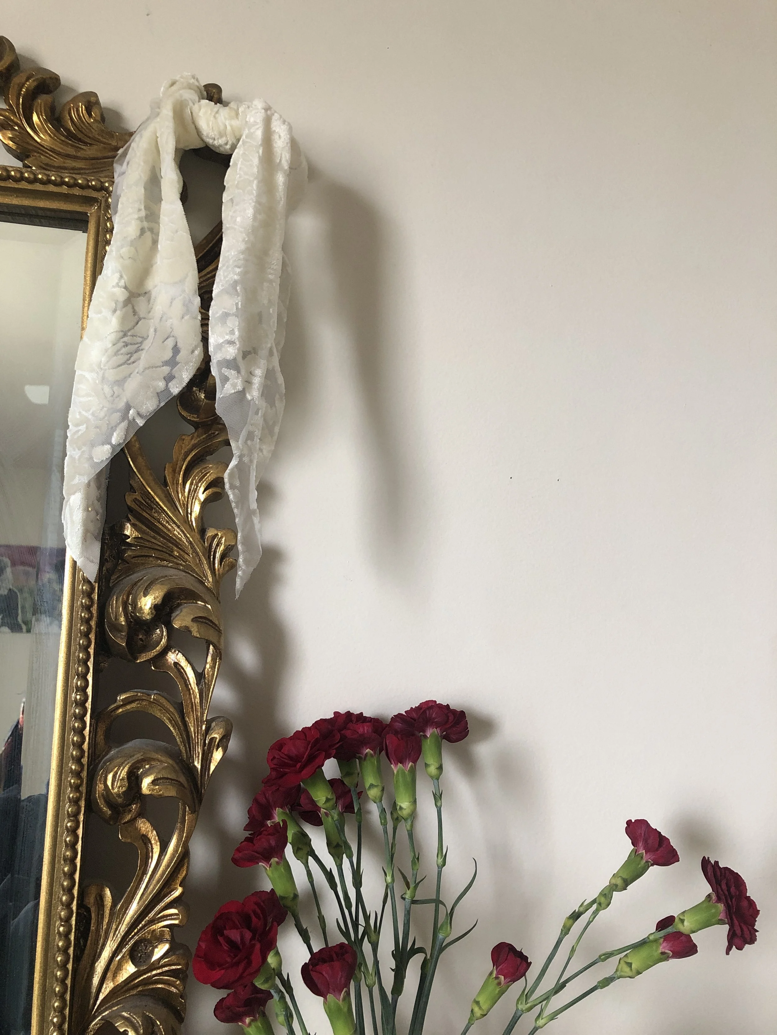 Close-up of a bouquet of red carnations in front of a white wall, with a gold ornate mirror frame and a white lace ribbon hanging from it.