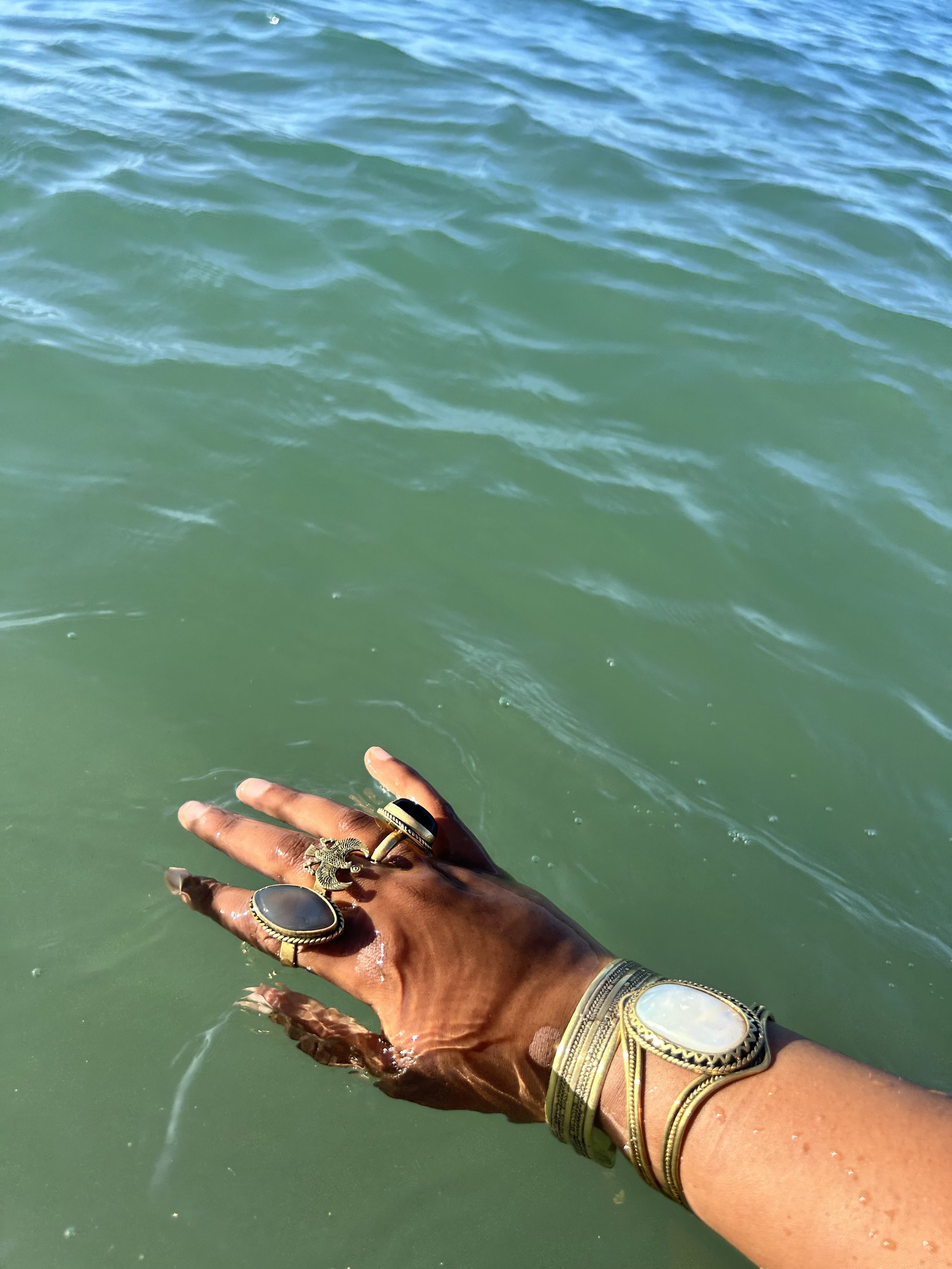 A person with dark skin wearing multiple rings and bracelets on their hand and wrist, reaching into a body of water with greenish color and small waves, possibly a lake or ocean.