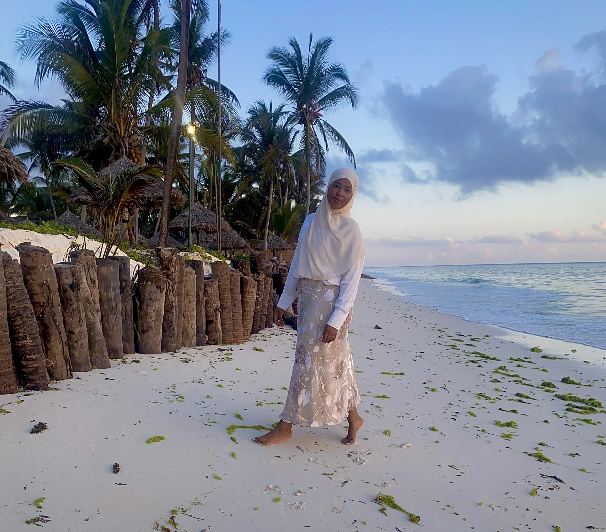 A woman in white hijab and long skirt standing barefoot on sandy beach near turquoise water with palm trees and thatched huts in the background during sunset.