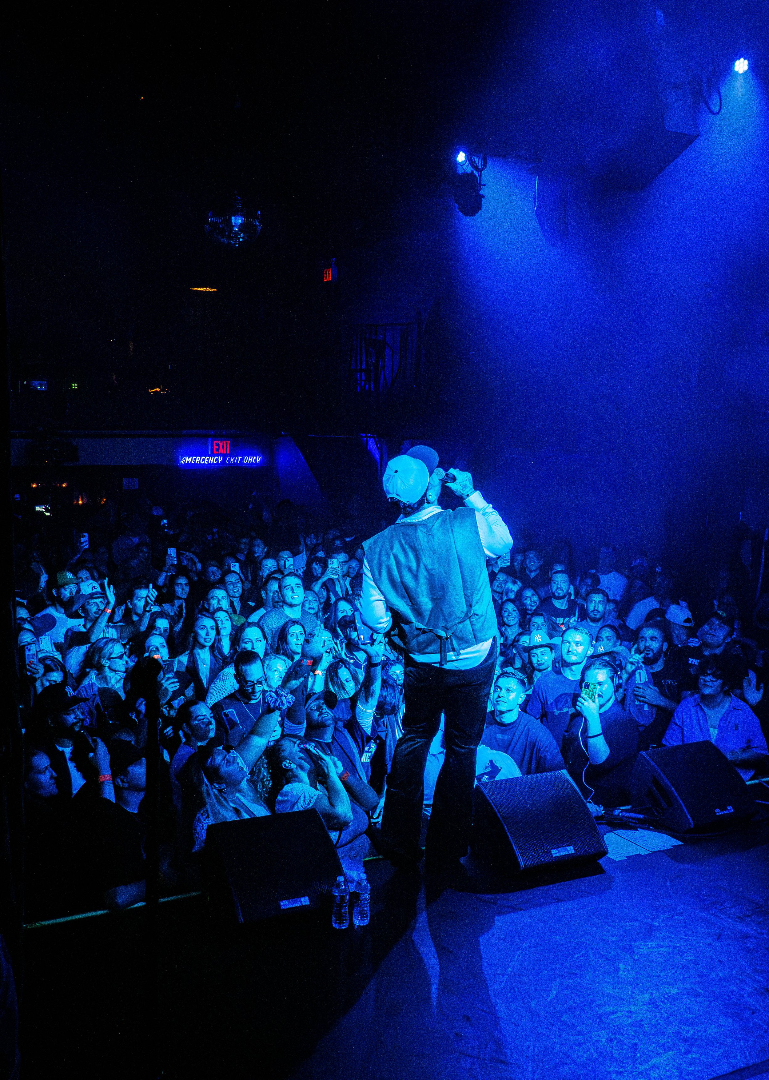 Performer on stage singing to an audience in a dark, blue-lit concert venue with many fans taking photos.