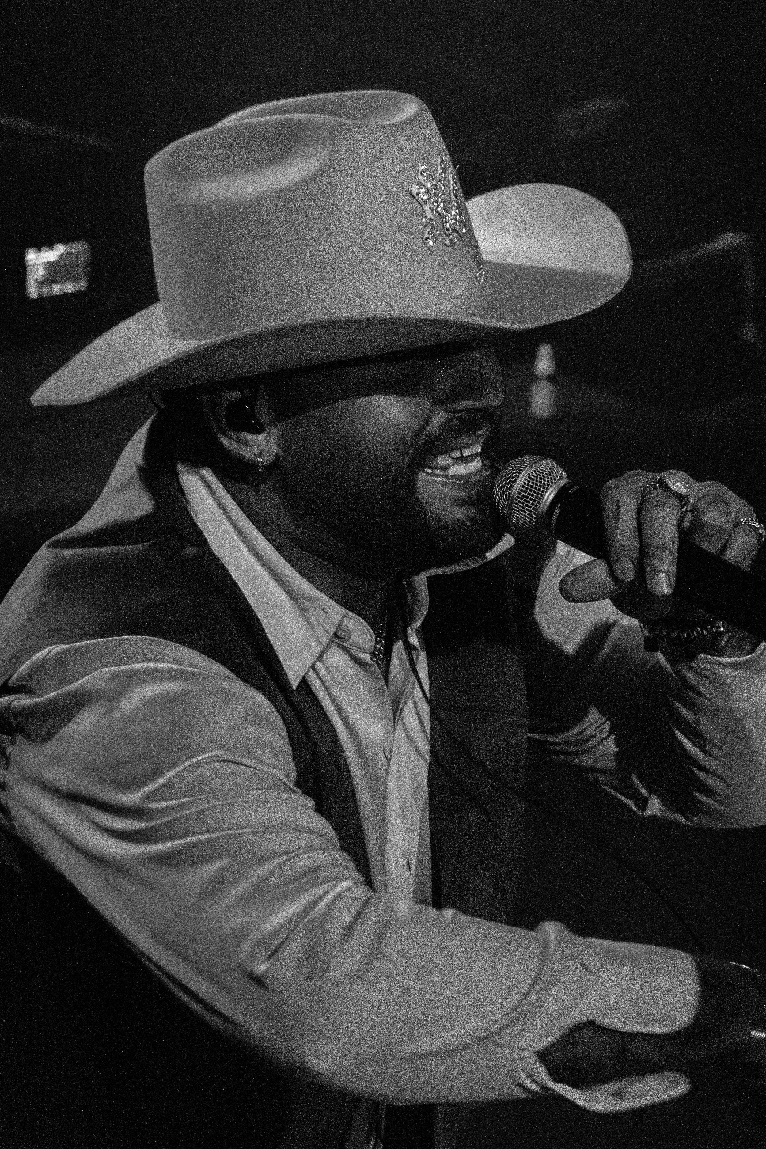 A man wearing a cowboy hat with a star badge, smiling, singing into a microphone in a black and white photo.