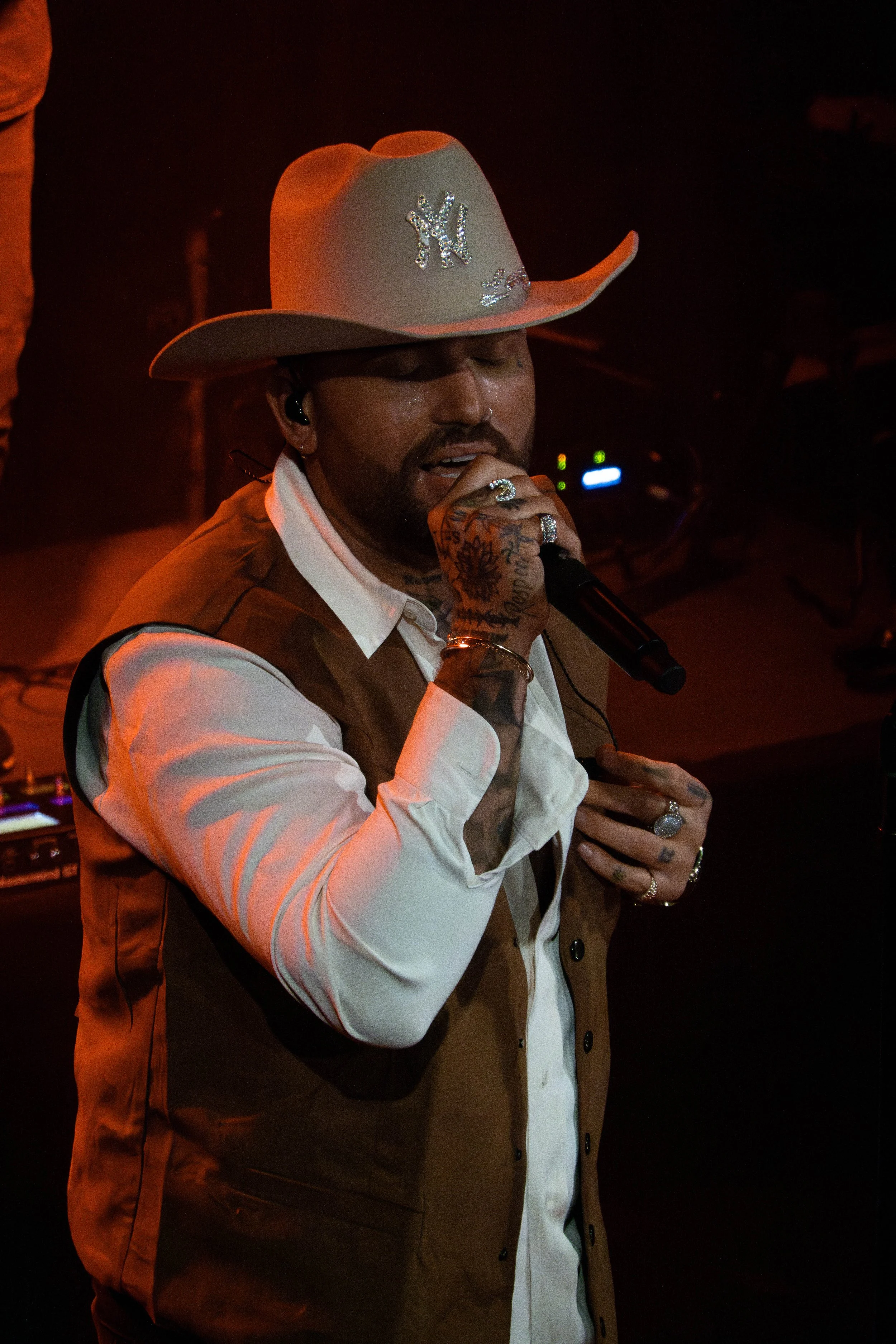 A man wearing a white cowboy hat with rhinestone embellishments, a white button-up shirt, and a brown vest, singing into a microphone on stage with dim, reddish lighting.