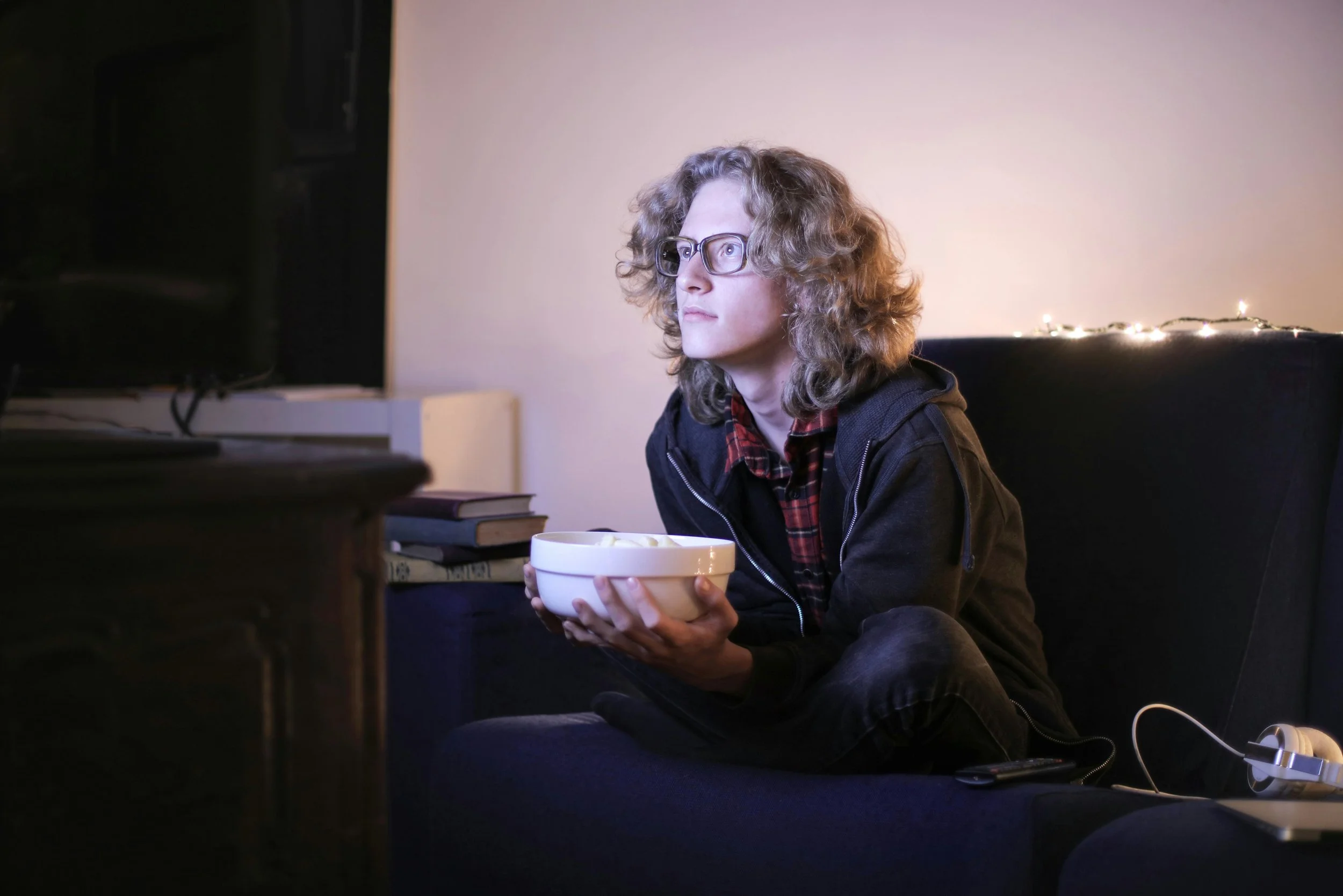 An androgynous, white adult with curly hair is wearing a flannel and a hoodie while holding a snack bowl and watching television.