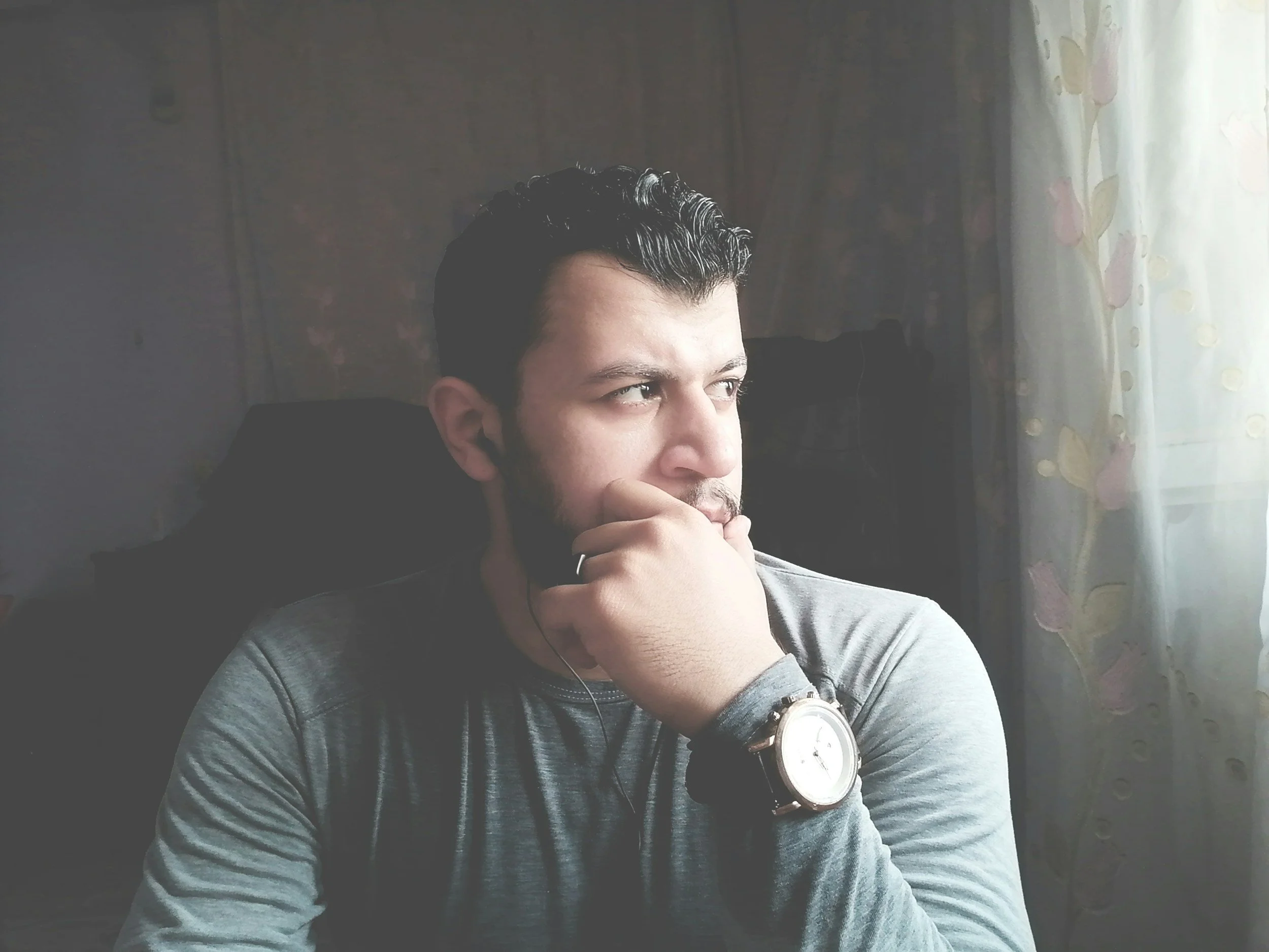 A man sitting indoors near a window with sheer curtains, looking thoughtfully to the right. He has dark hair, a beard, wears a watch on his left wrist, and has earbuds in his ears.