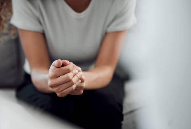 Close-up of woman sitting with hands clasped, wearing a light gray T-shirt and black pants, indoors.