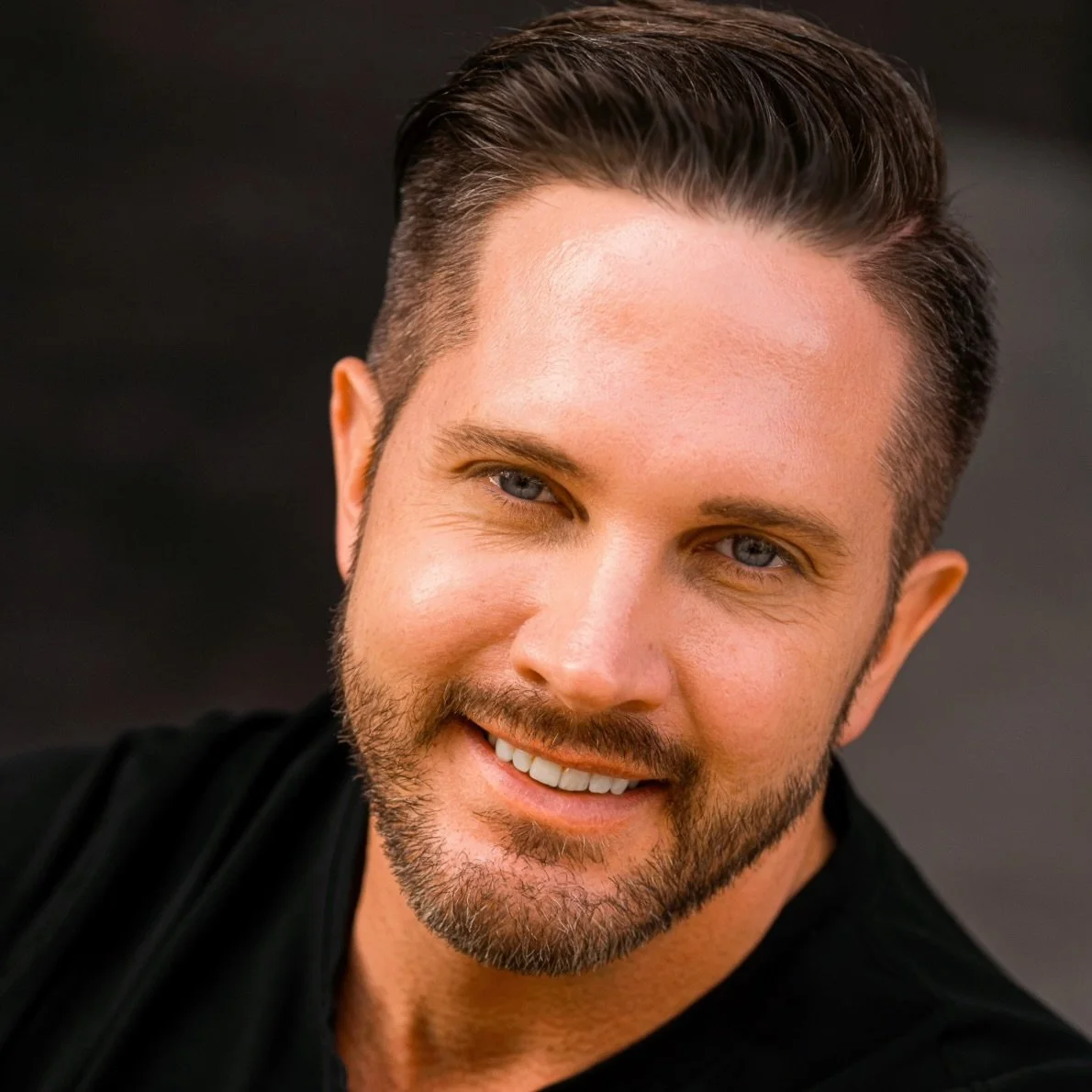 Close-up portrait of a smiling man with light skin, short brown hair, blue eyes, and a beard, wearing a black shirt, against a dark background.