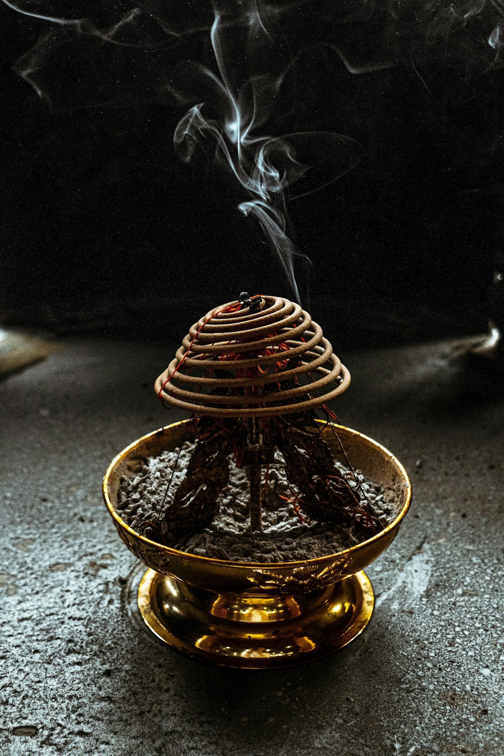 A brass incense burner with spiral coil incense emitting smoke, placed on a gray surface.