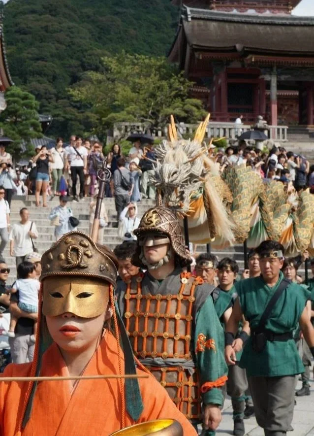 People participating in a traditional Japanese festival parade, wearing historical costumes including masks and armor, with a Japanese temple and green mountainous landscape in the background.