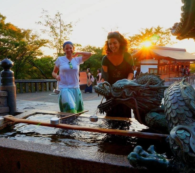 Two women smiling at a water fountain featuring a dragon sculpture at sunset, with traditional Asian-style architecture and trees in the background.