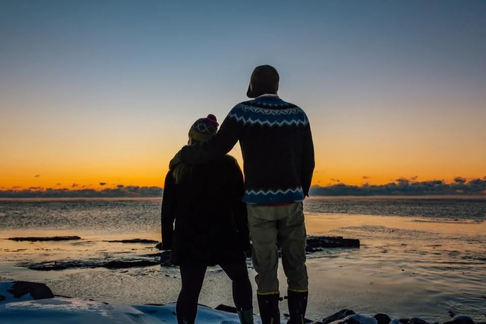 Two people, wearing dark winter clothing and knit hats, are watching fireworks in the night sky.