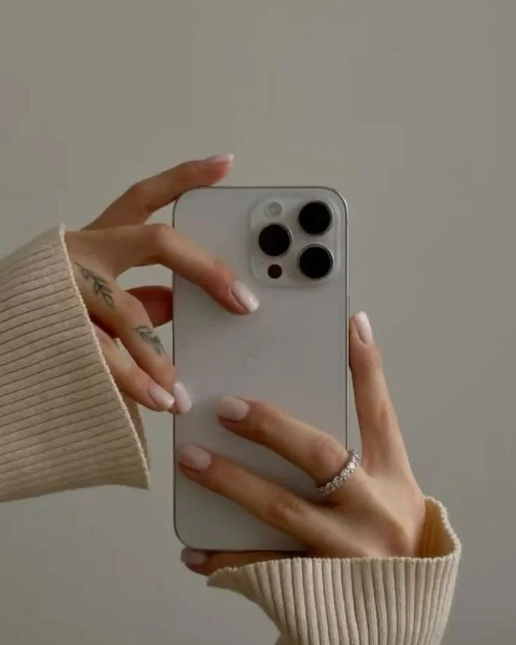 Person taking a mirror selfie with a white smartphone, wearing a beige sweater and a ring