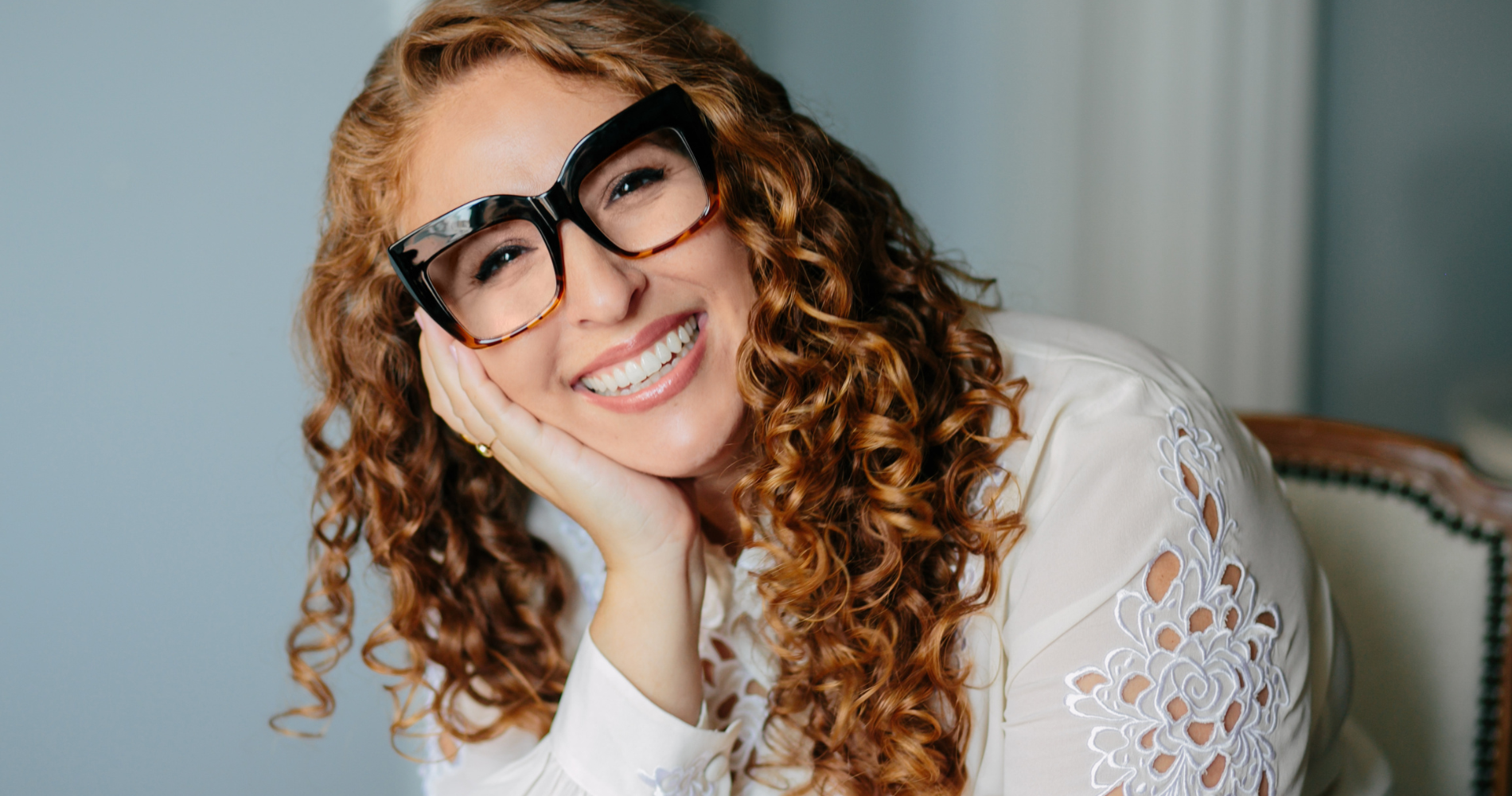 A woman with curly red hair wearing large black glasses and a white embroidered blouse, smiling and resting her face on her hand.