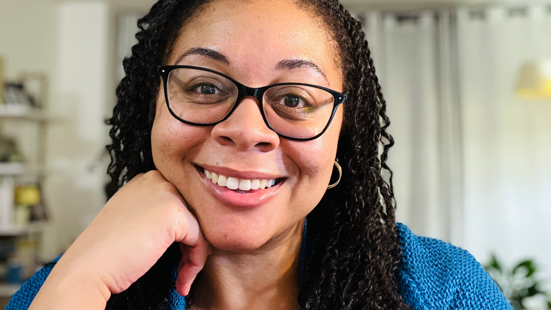 Close-up of a smiling woman with glasses and curly hair, wearing a blue sweater, sitting indoors with a blurred background.