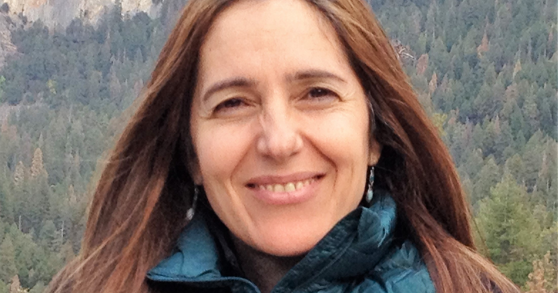 Close-up of a woman smiling outdoors, wearing a blue jacket and earrings, with a forested mountain in the background.