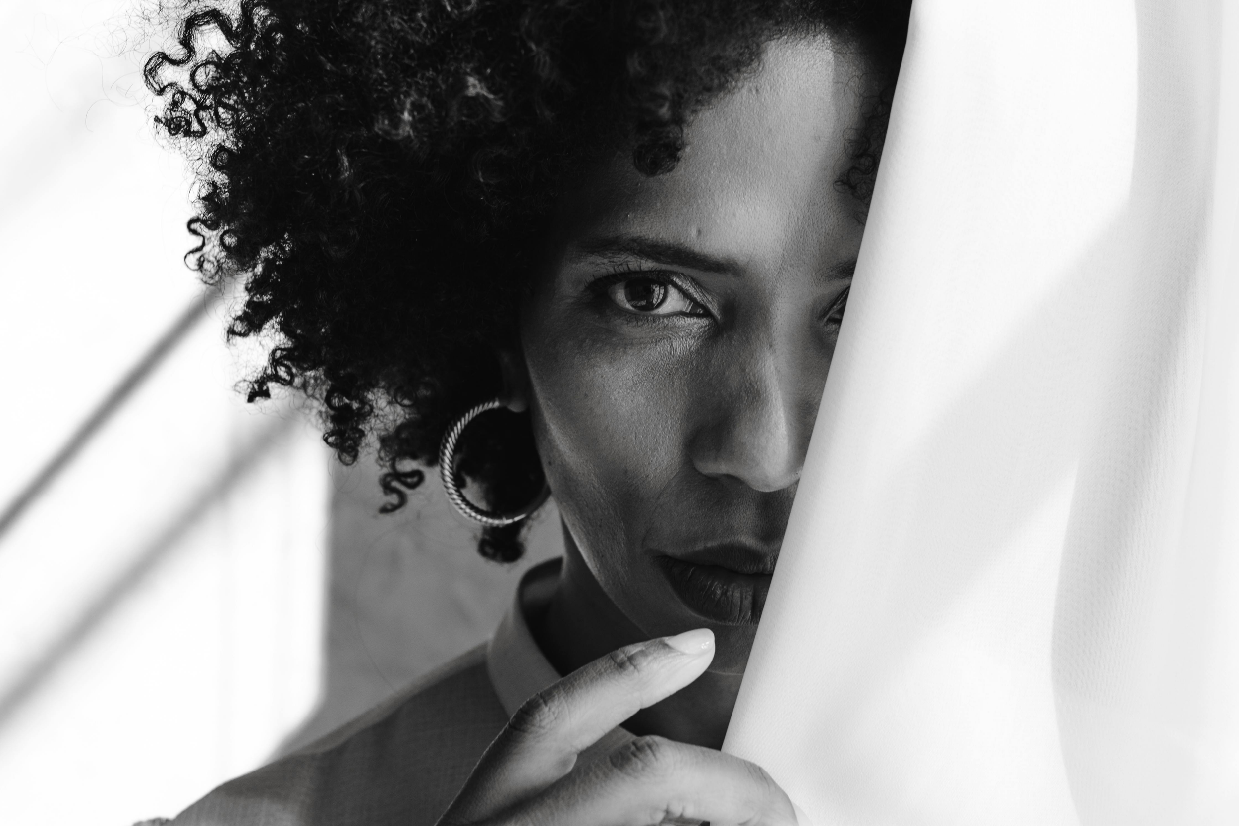 A close-up black and white photograph of a woman with curly hair looking over a partially open white door.