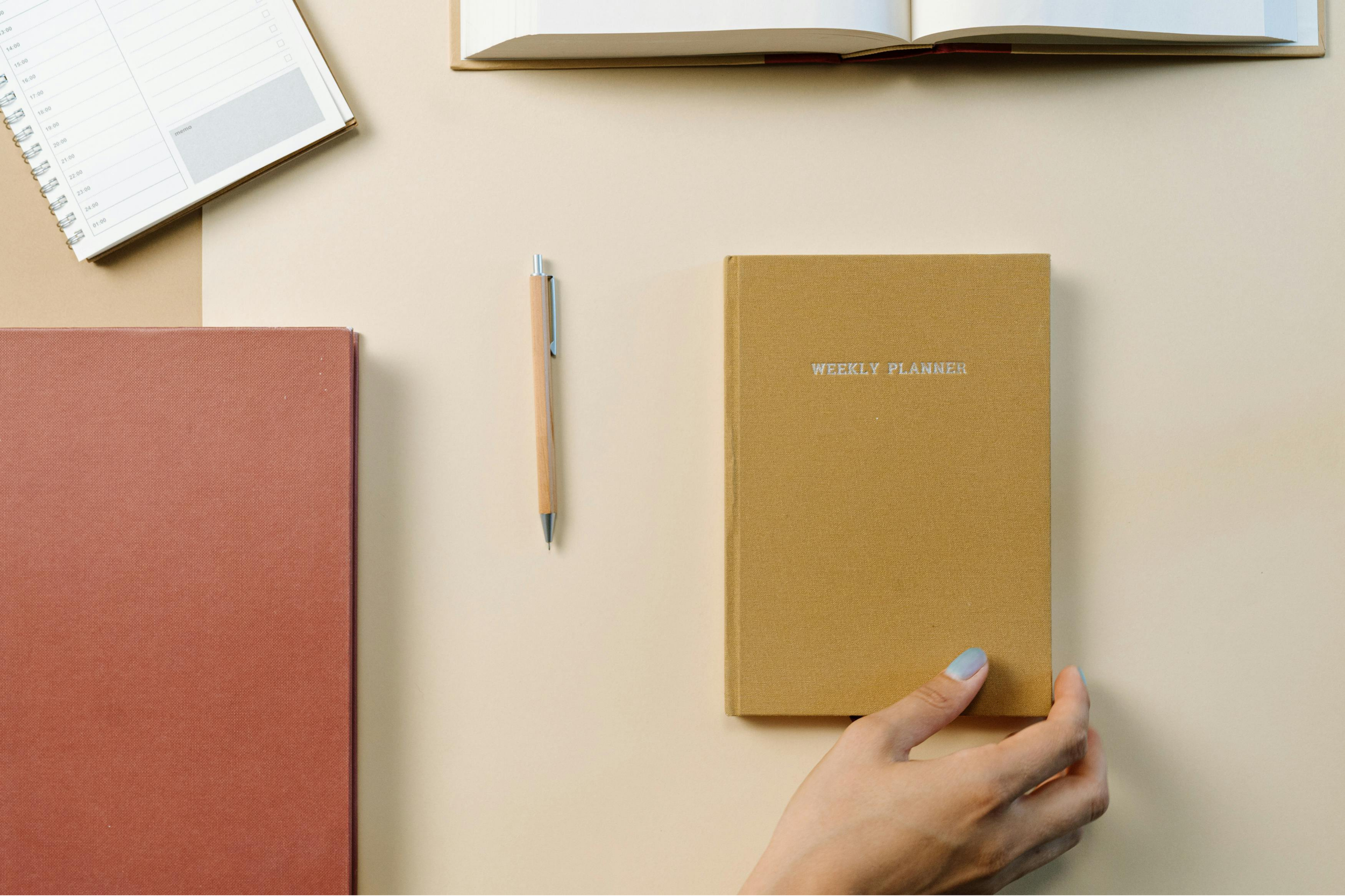 A person's hand holding a gold-colored weekly planner on a beige surface, with an open book, a pink notebook, a pen, and a calendar planner nearby.