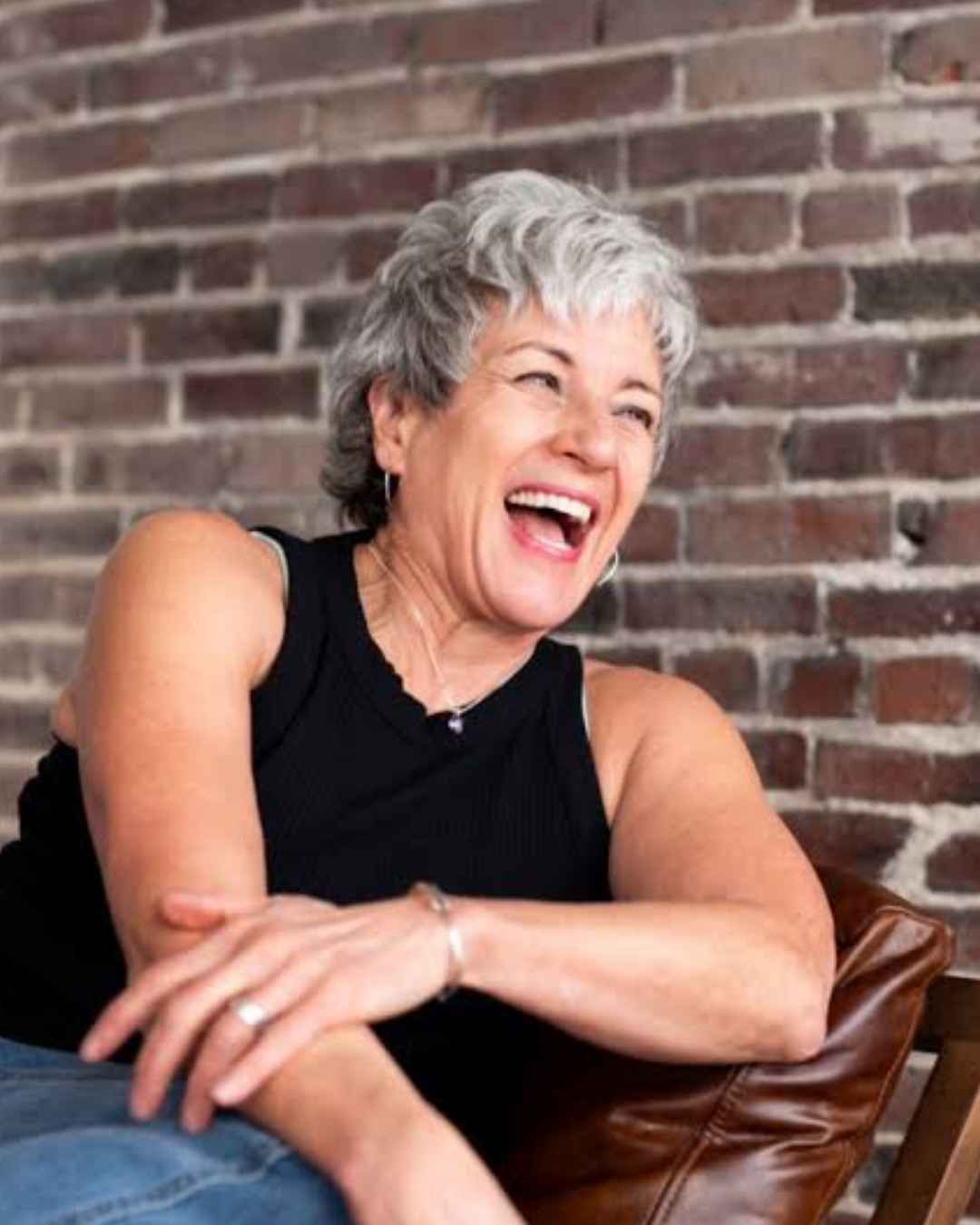 Older woman with short gray hair laughing, sitting against a brick wall with arms crossed and wearing a black sleeveless top.