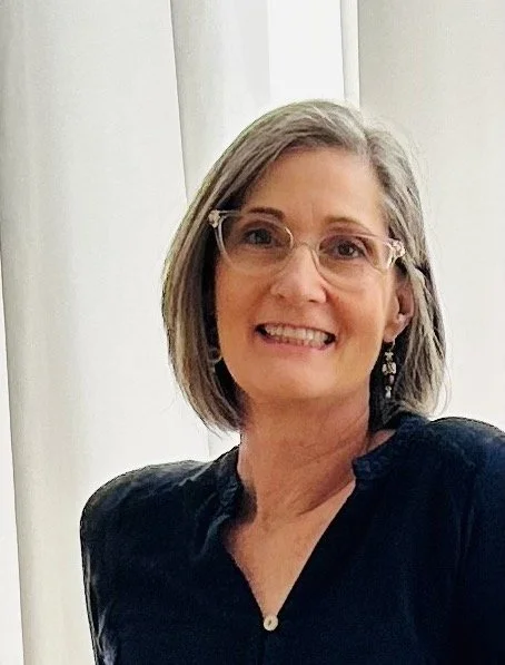 Professional health and wellness coach with short gray hair and glasses, wearing earrings and a black top, seated indoors in front of white curtains.