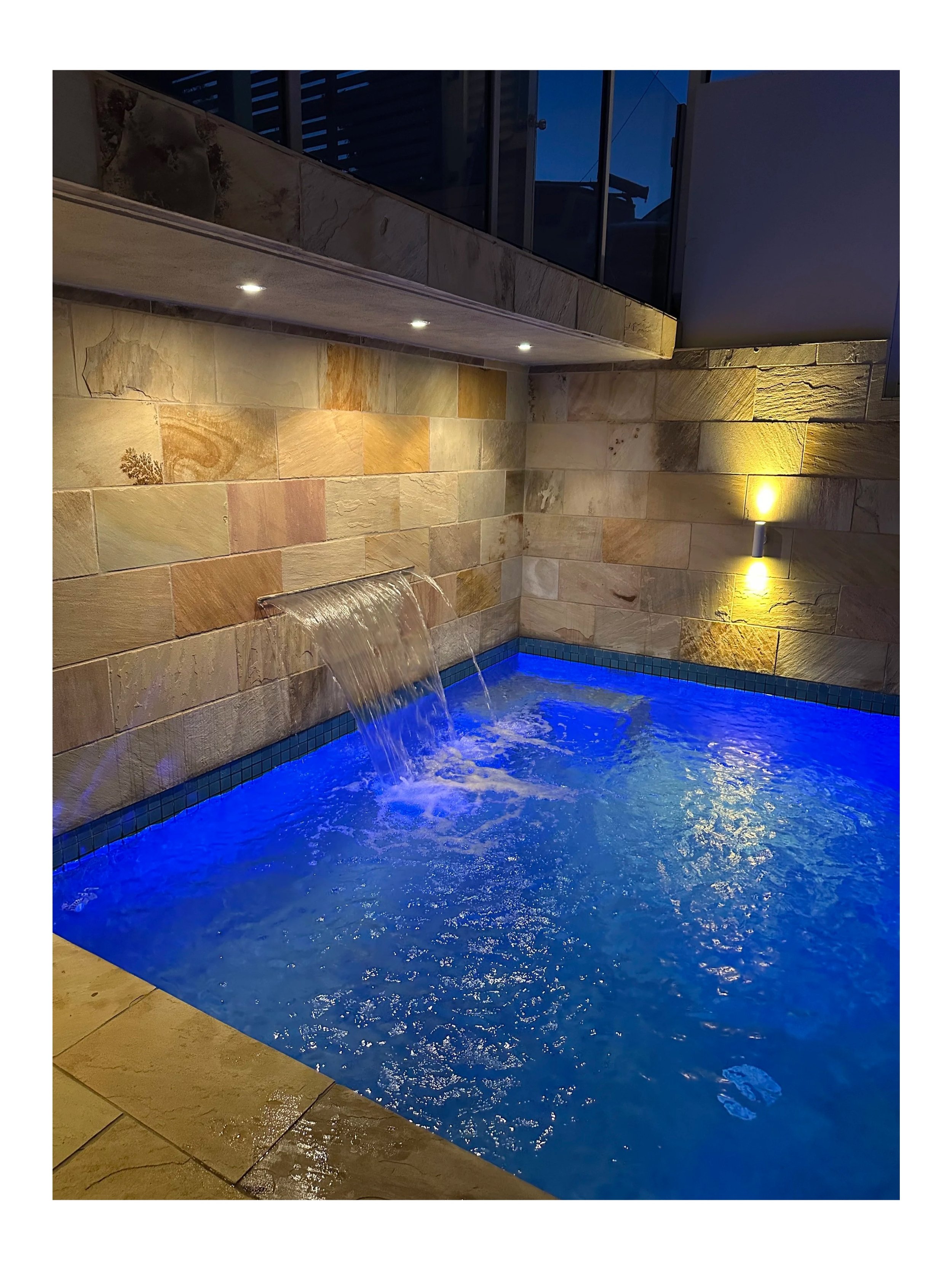 Indoor pool at night with blue lighting, waterfall feature, stone wall, and wall-mounted lights.
