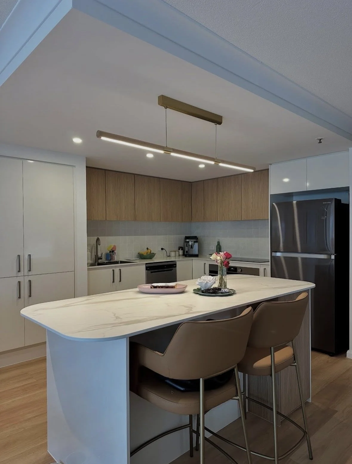 Modern kitchen with a white marble island, beige chairs, wooden upper cabinets, white lower cabinets, stainless steel refrigerator, and a minimalist pendant light.