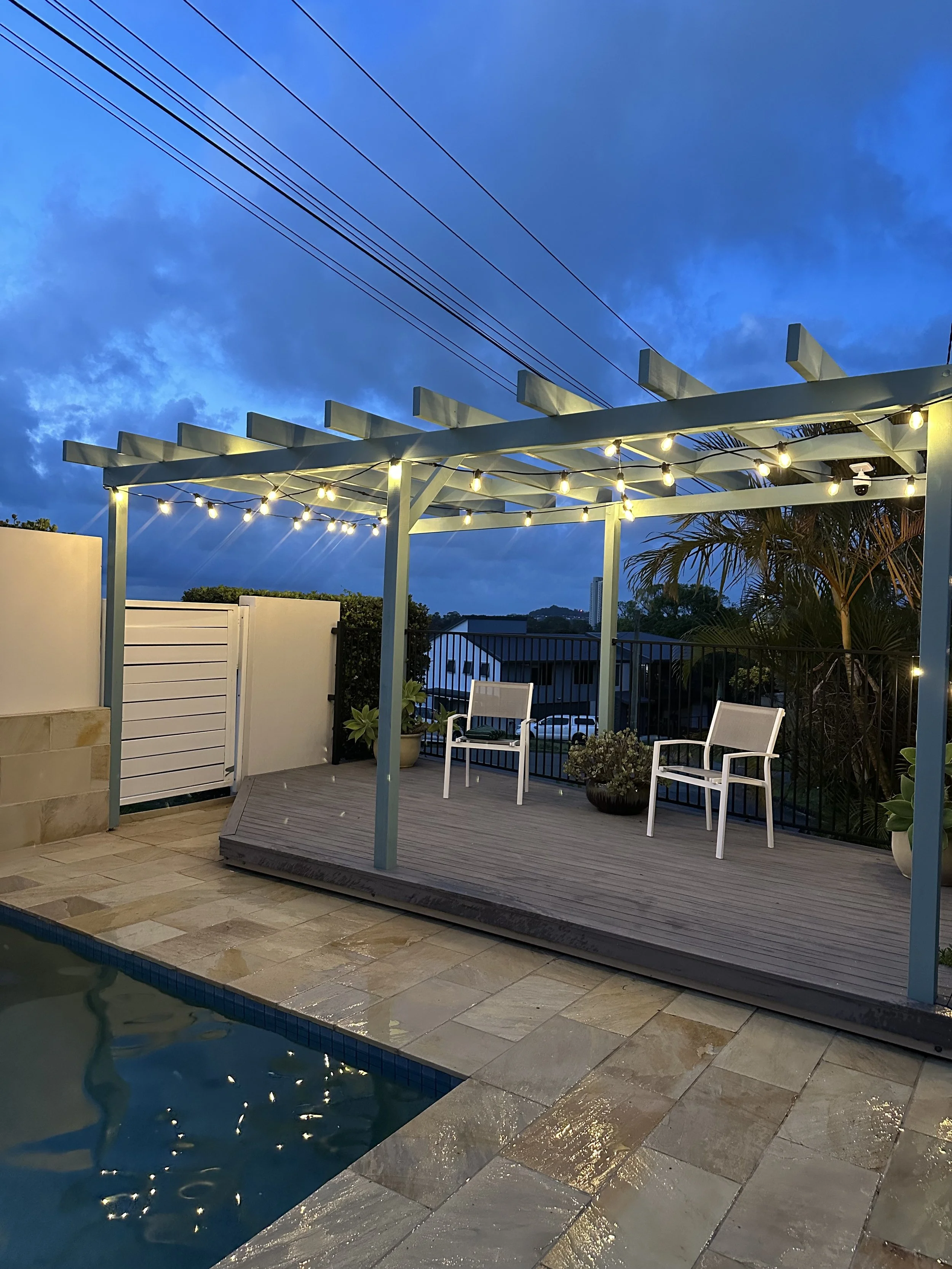 Backyard patio with a small pool, wooden deck, two white chairs, potted plants, and string lights, during dusk with a cloudy sky.