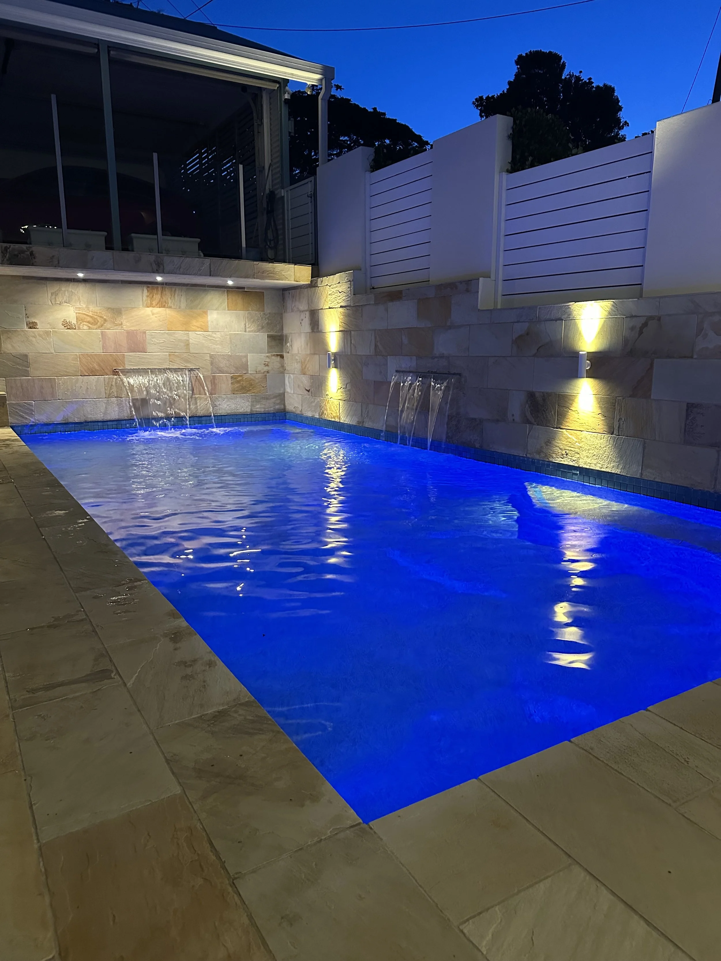 A nighttime view of a small, illuminated outdoor swimming pool with water features and blue LED lighting, surrounded by a stone-tiled deck and a modern white fence.