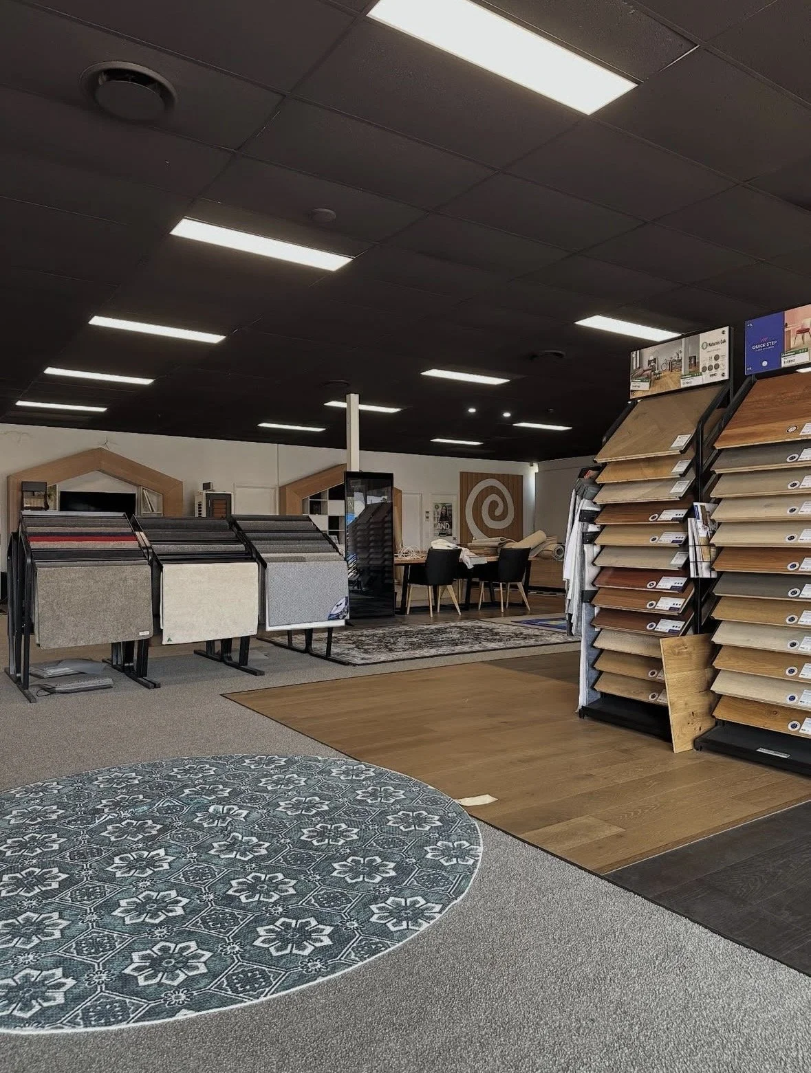 Interior of a flooring store with display samples of carpets, hardwood, and laminate flooring, and a seating area in the background.