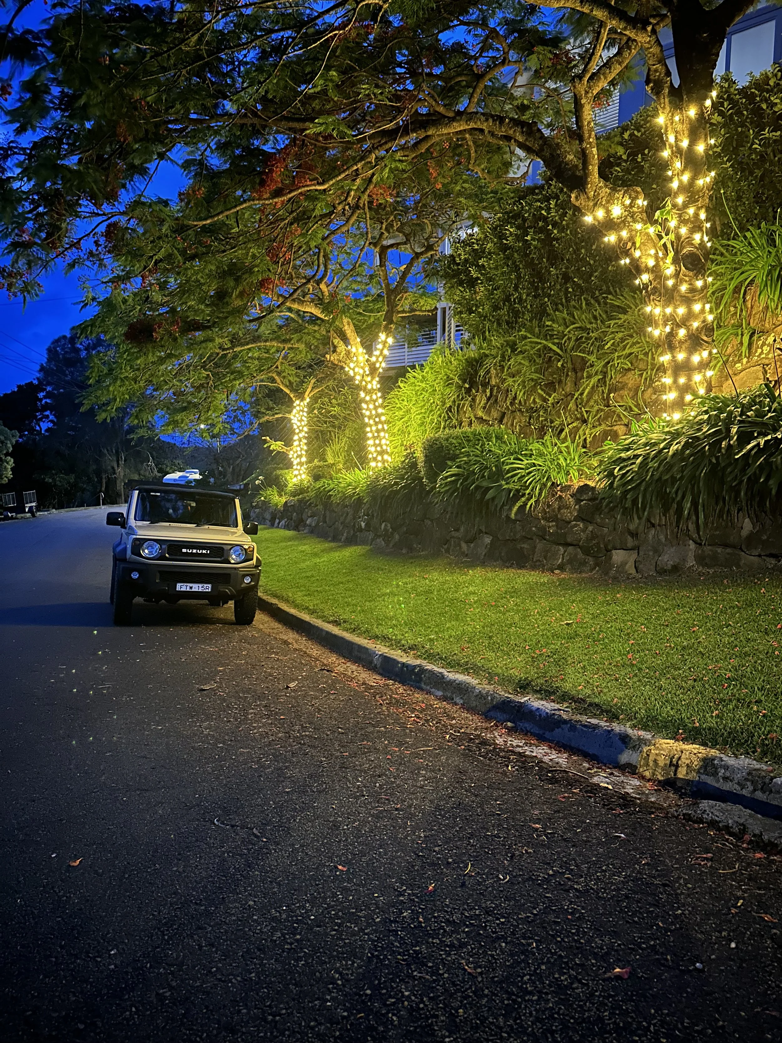 A black Suzuki Jimny parked on the side of a street at dusk. Trees with string lights wrapped around the trunks and lit leaves are illuminated by nearby lights, with greenery on a rock wall in the background.