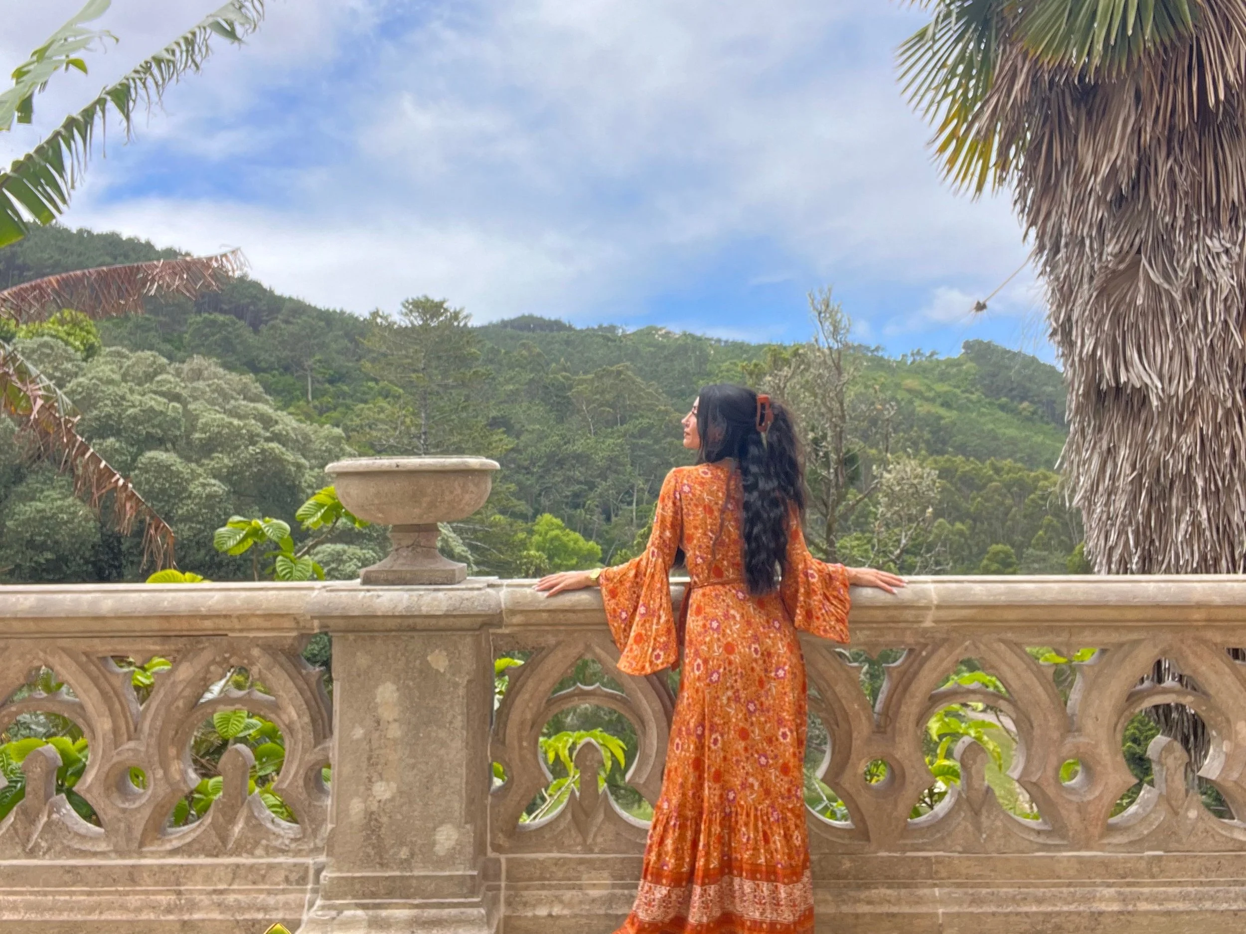 A woman in an orange patterned dress with long dark hair stands on a stone balcony, overlooking a lush green forest and hills under a blue sky with light clouds.