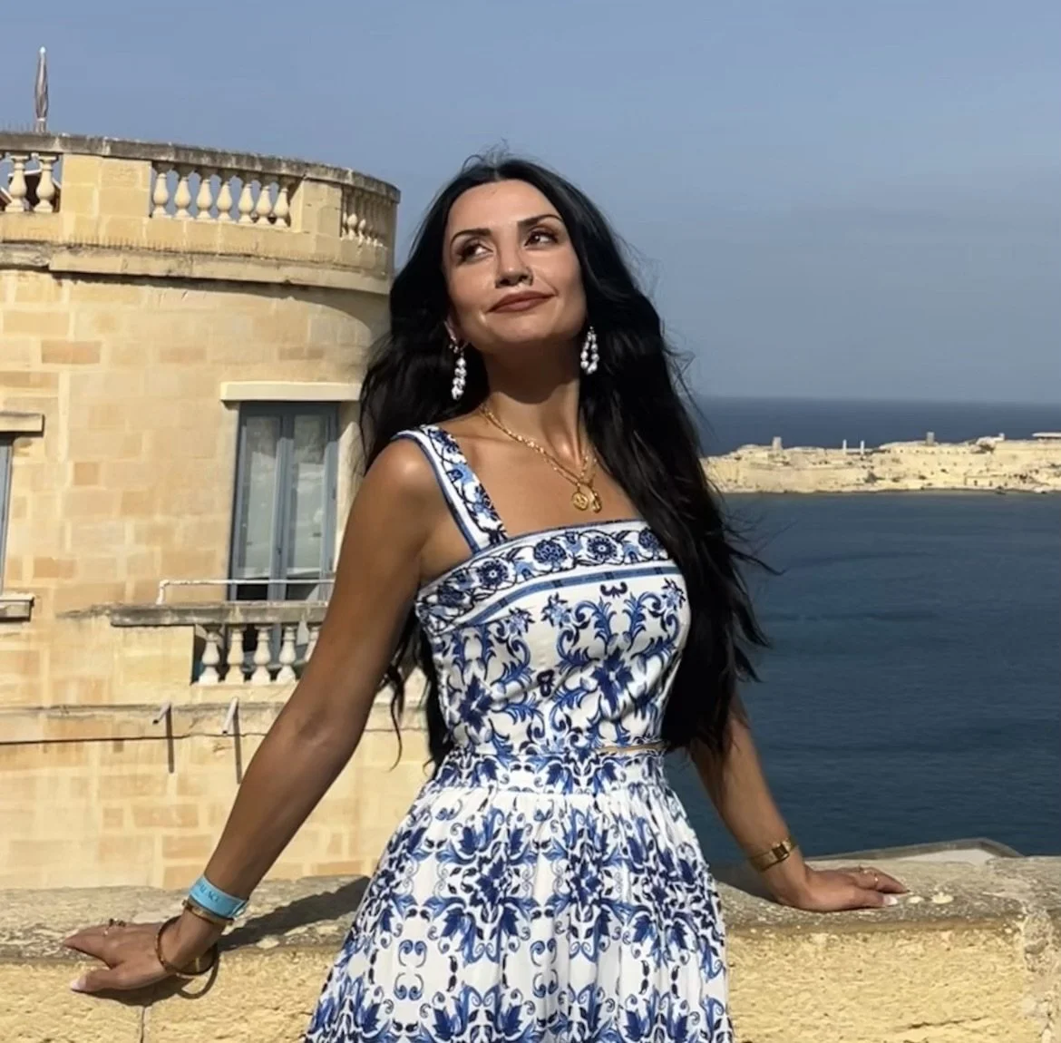 A woman in a blue and white patterned dress standing on a balcony overlooking the sea and a historic building, with clear blue sky in the background.