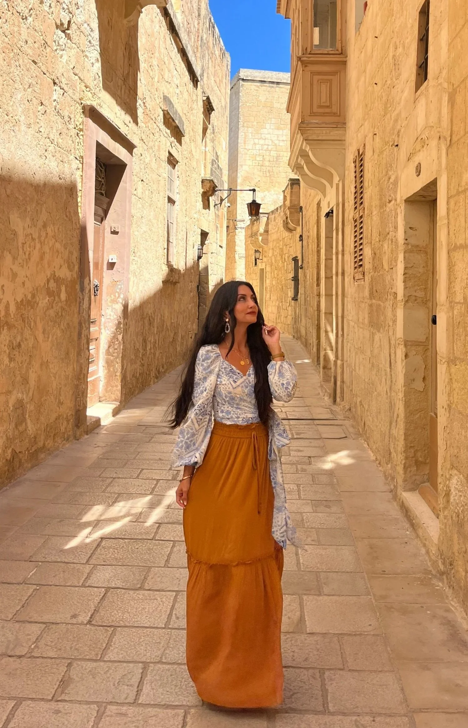 A woman with long dark hair wearing a light-colored blouse and an orange skirt walking down a narrow stone-paved alleyway with beige stone buildings and blue sky overhead.