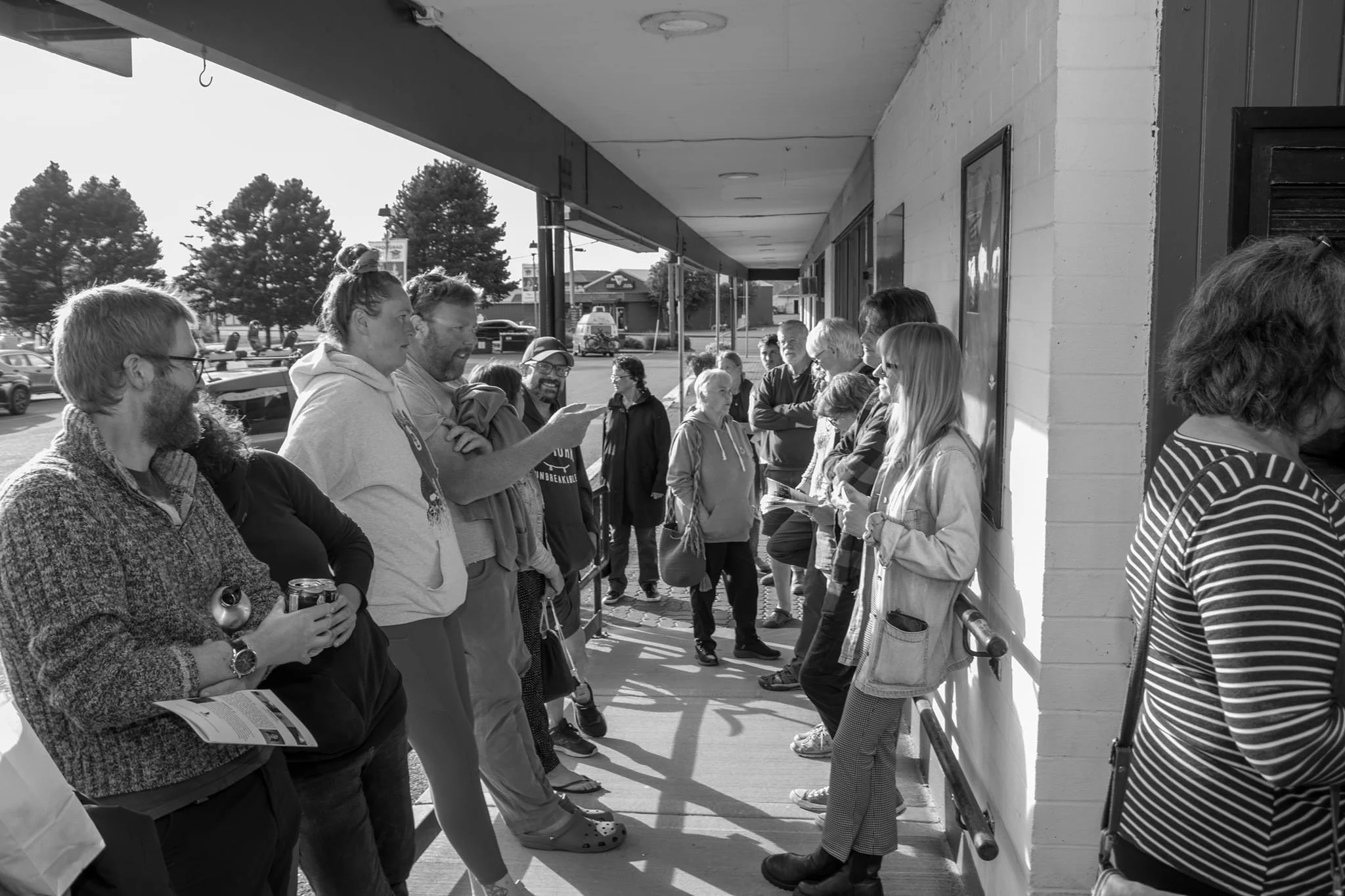 Group of people waiting in line outside a building on a sidewalk, some are engaged in conversation, and a few are holding papers or beverages.